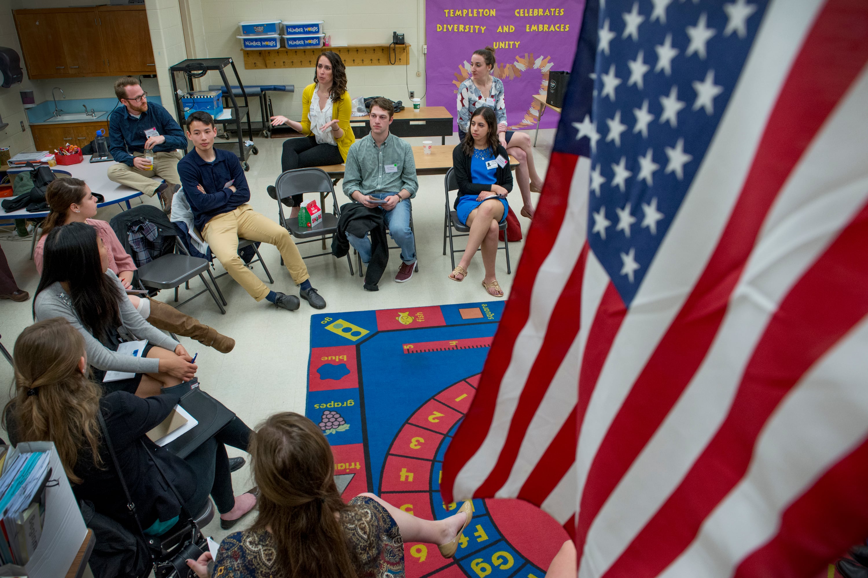 A large American flag hangs in the right foreground while, in the background, people sit in chairs around a colorful rug in a classroom.