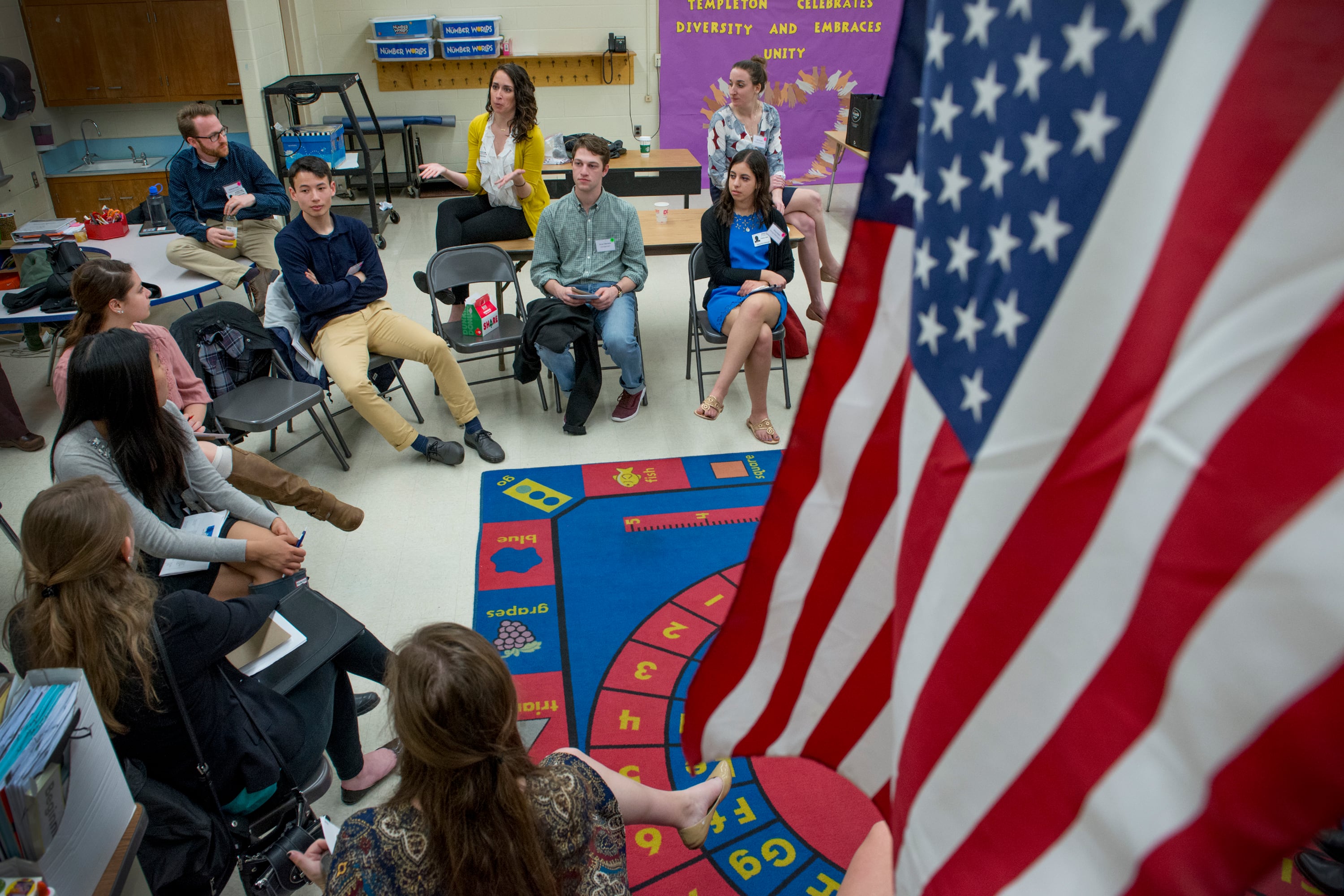 A large American flag hangs in the right foreground while, in the background, people sit in chairs around a colorful rug in a classroom.