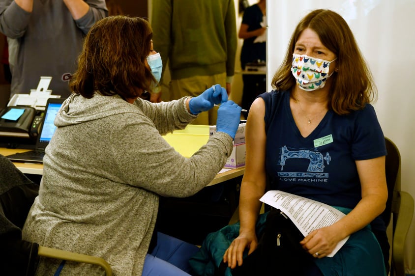 A medical worker administers a COVID-19 vaccine in the right arm of a school nurse as she sits.