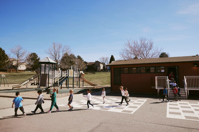 A line of students walk across a concrete playground with a play set in the background next to a small school building. A blue sky in the background.