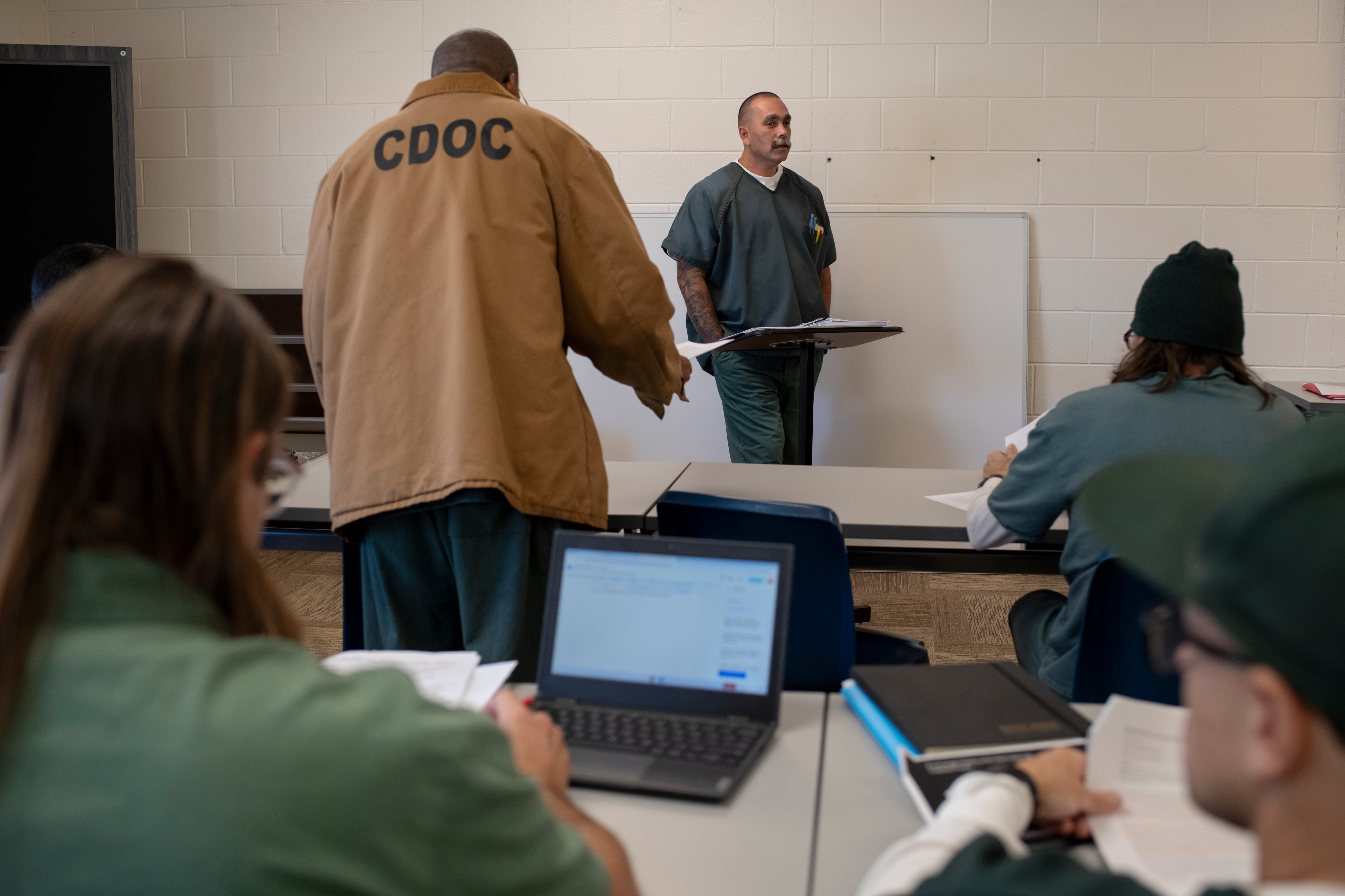 A man wearing a green jump suit stands at the front of a class with several others sitting at desks and one person standing with a tan jacket that reads "CDOC". A white brick wall in the background.