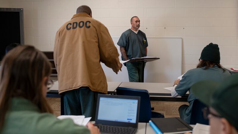 A man wearing a green jump suit stands at the front of a class with several others sitting at desks and one person standing with a tan jacket that reads "CDOC". A white brick wall in the background.