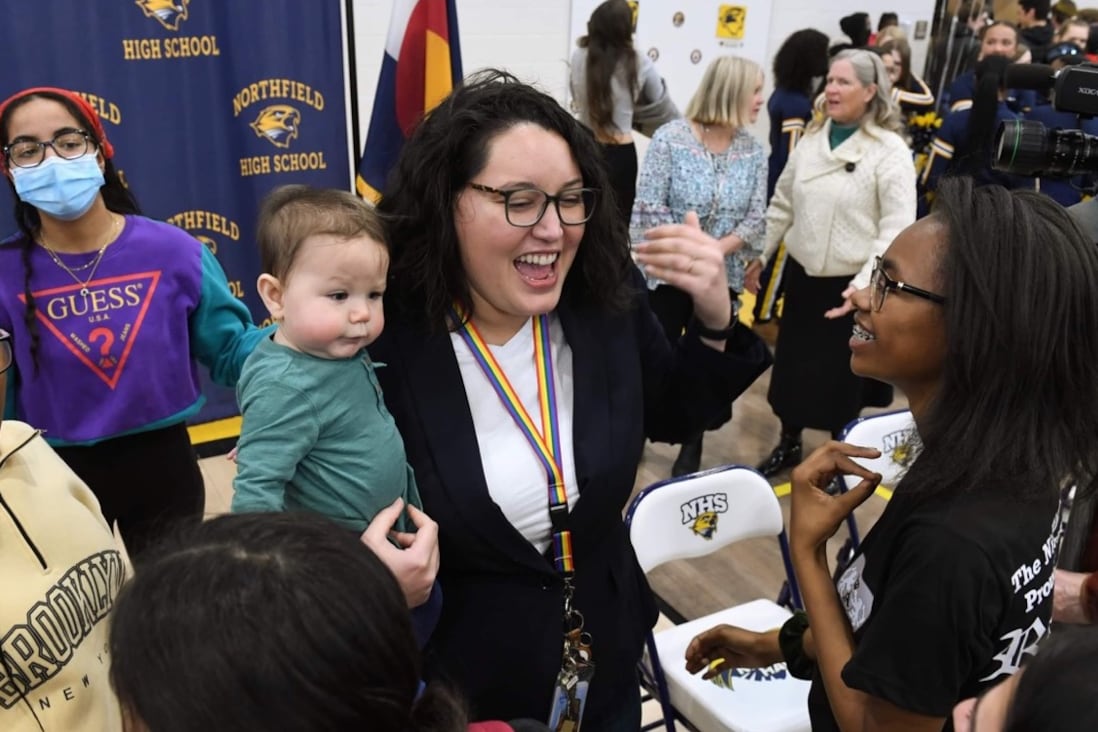 A high school teacher holds a baby while she talks to a student in a crowded gym.