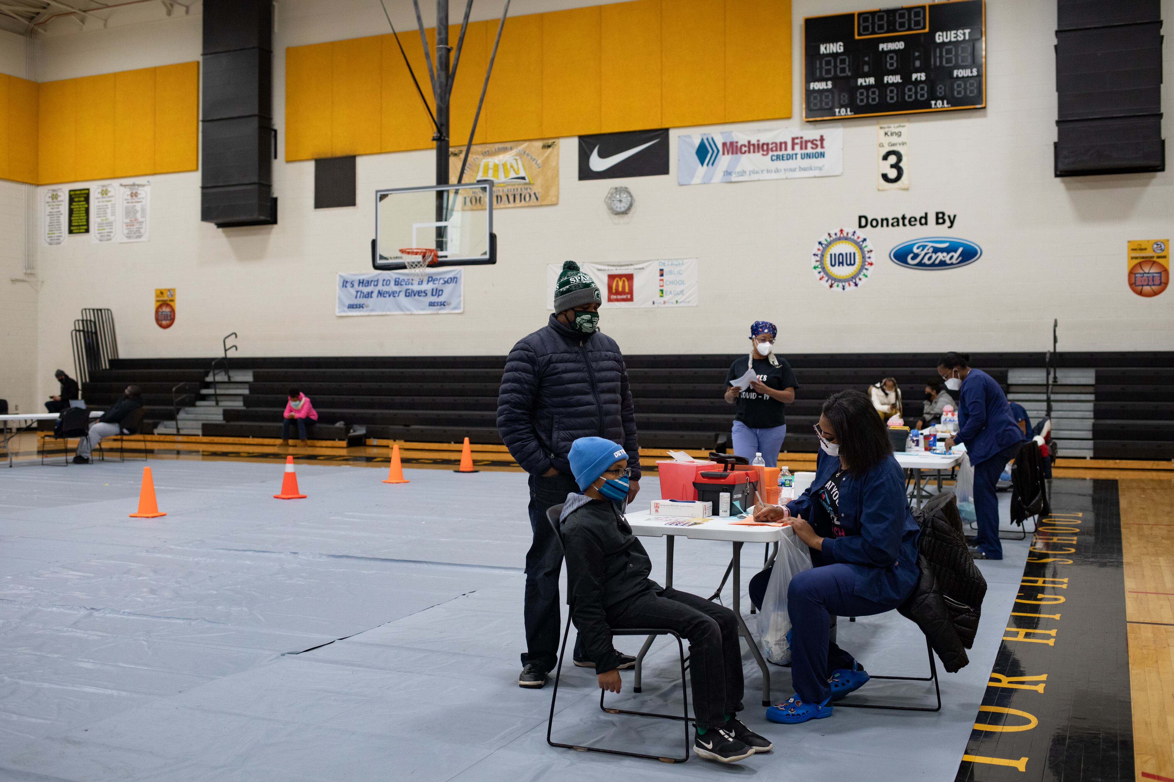 An adult stands beside a sitting child while a nurse writes down information on a table.