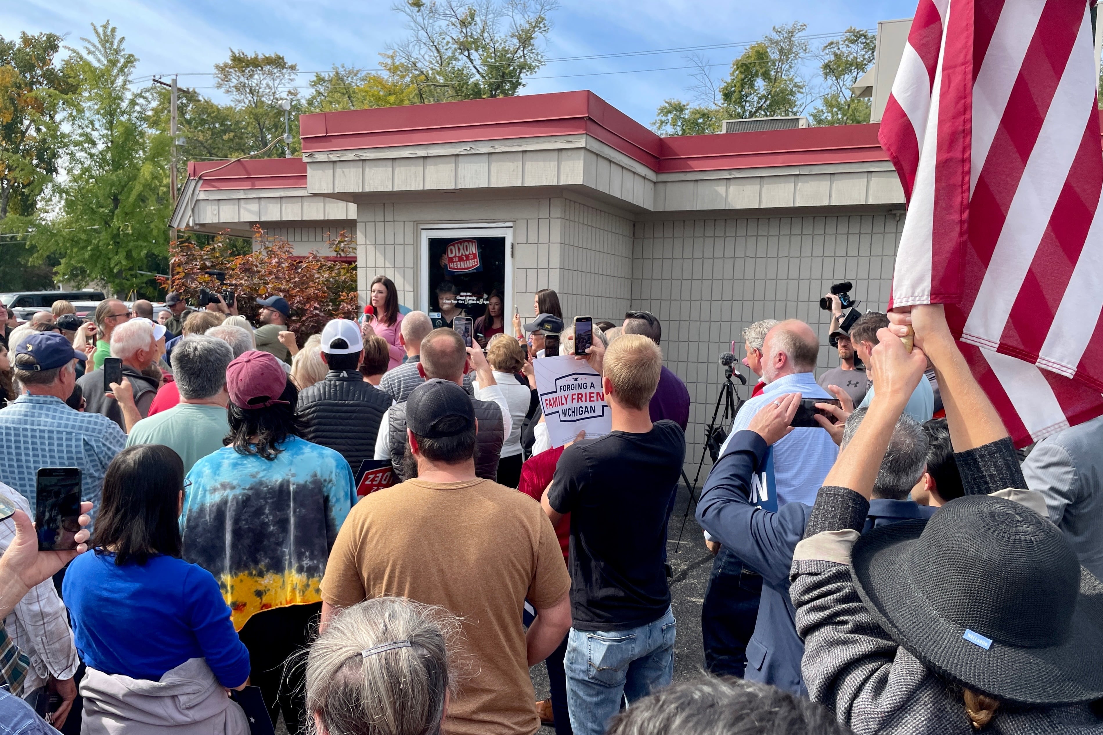 A crowd of supporters listen to Tudor Dixon, who is speaking into a microphone on the steps to the back entrance of a restaurant. Many are recording video on cell phones.