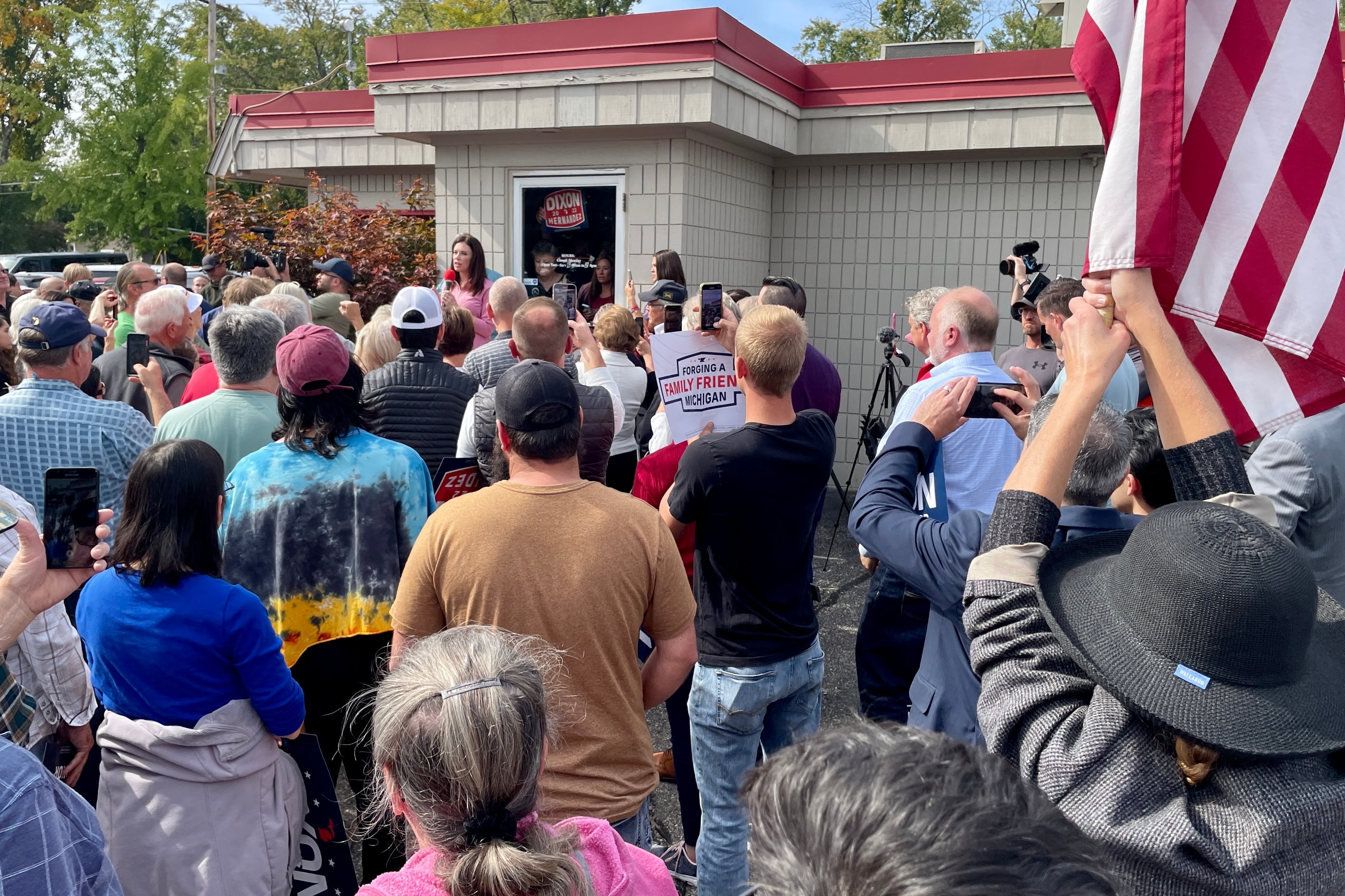 A crowd of supporters listen to Tudor Dixon, who is speaking into a microphone on the steps to the back entrance of a restaurant. Many are recording video on cell phones.