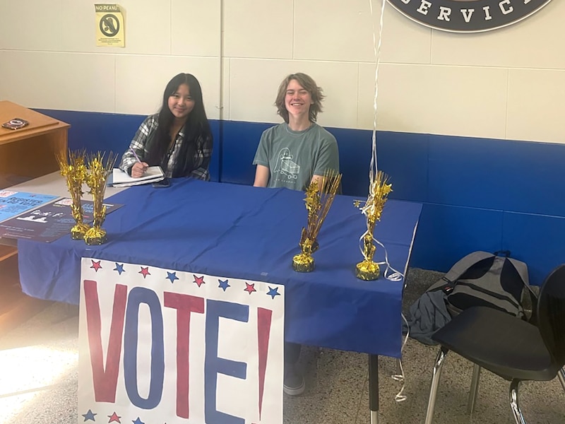 Two high school students sit behind a table with a large sign that says "VOTE!"