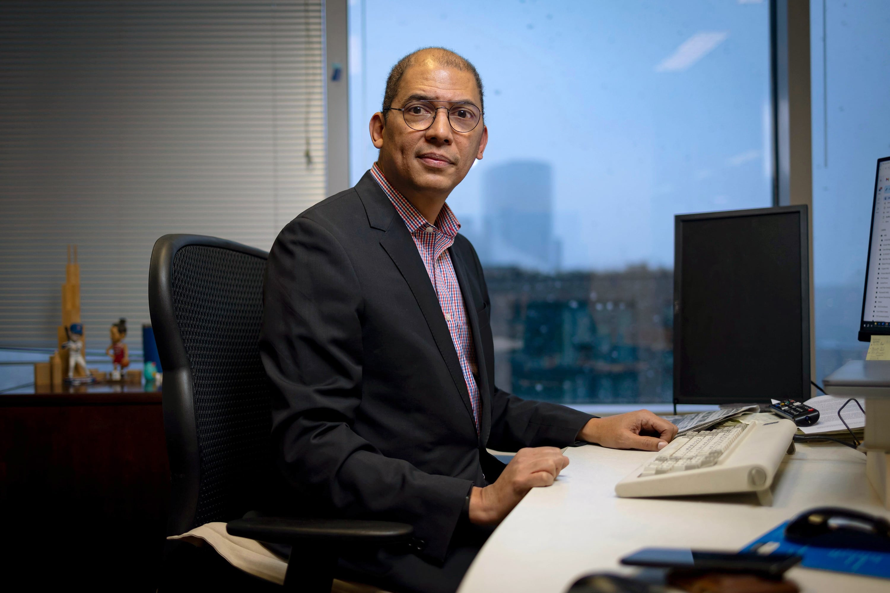 A man in a suit sits at a desk with a window in the background.