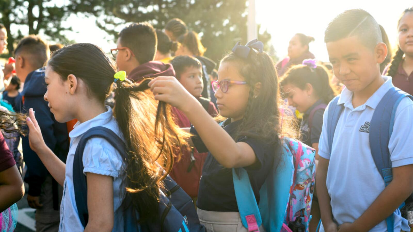 An elementary-age girl braids the hair of another girl as they stand amid a group of students wearing backpacks in 2018.