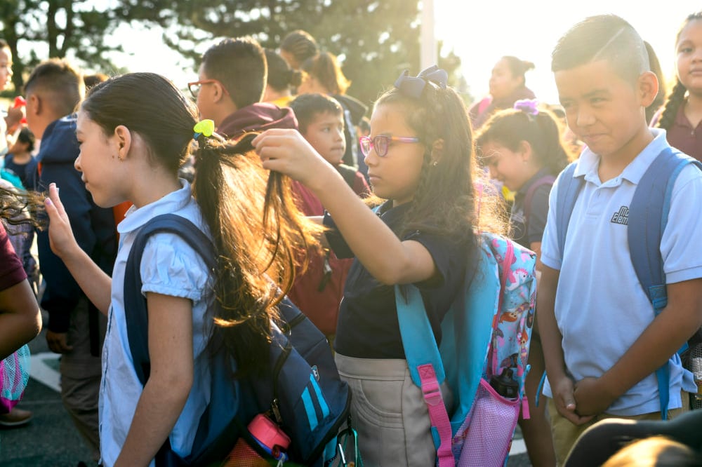 An elementary-age girl braids the hair of another girl as they stand amid a group of students wearing backpacks in 2018.