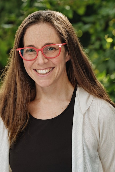 Headshot of a woman with long brown hair wearing red glasses, a black t-shirt, and a white button-down.