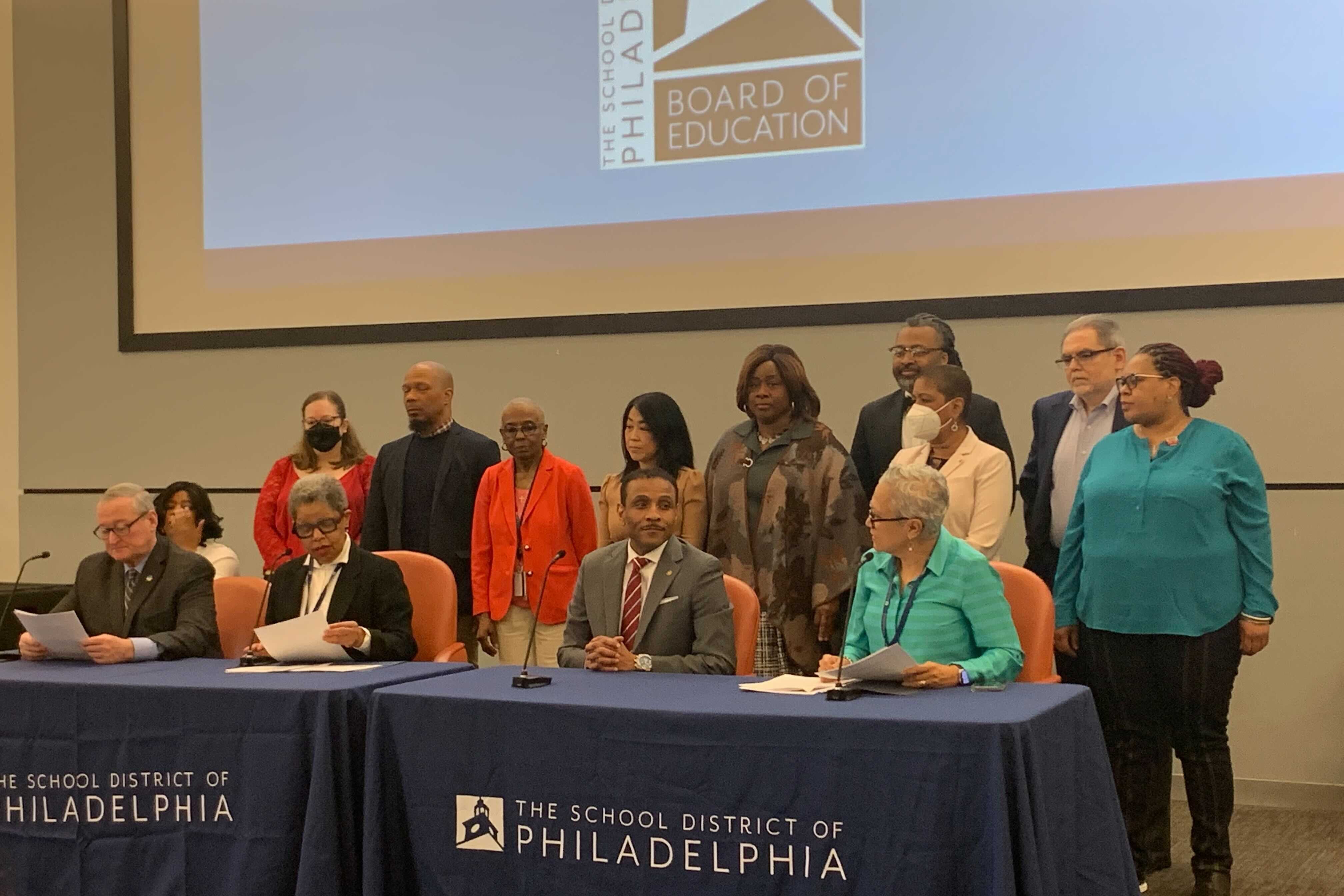 Four people sit at tables covered in blue cloth that says “The School District of Phildelphia.” There is a crowd of people behind them.