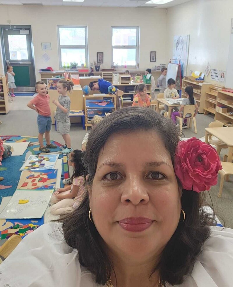 A woman with dark hair and a bright pink flower on the side of her hair is in the center of the image with her classroom of young children in the background. There's a colorful rug  on the floor and some windows in the back.