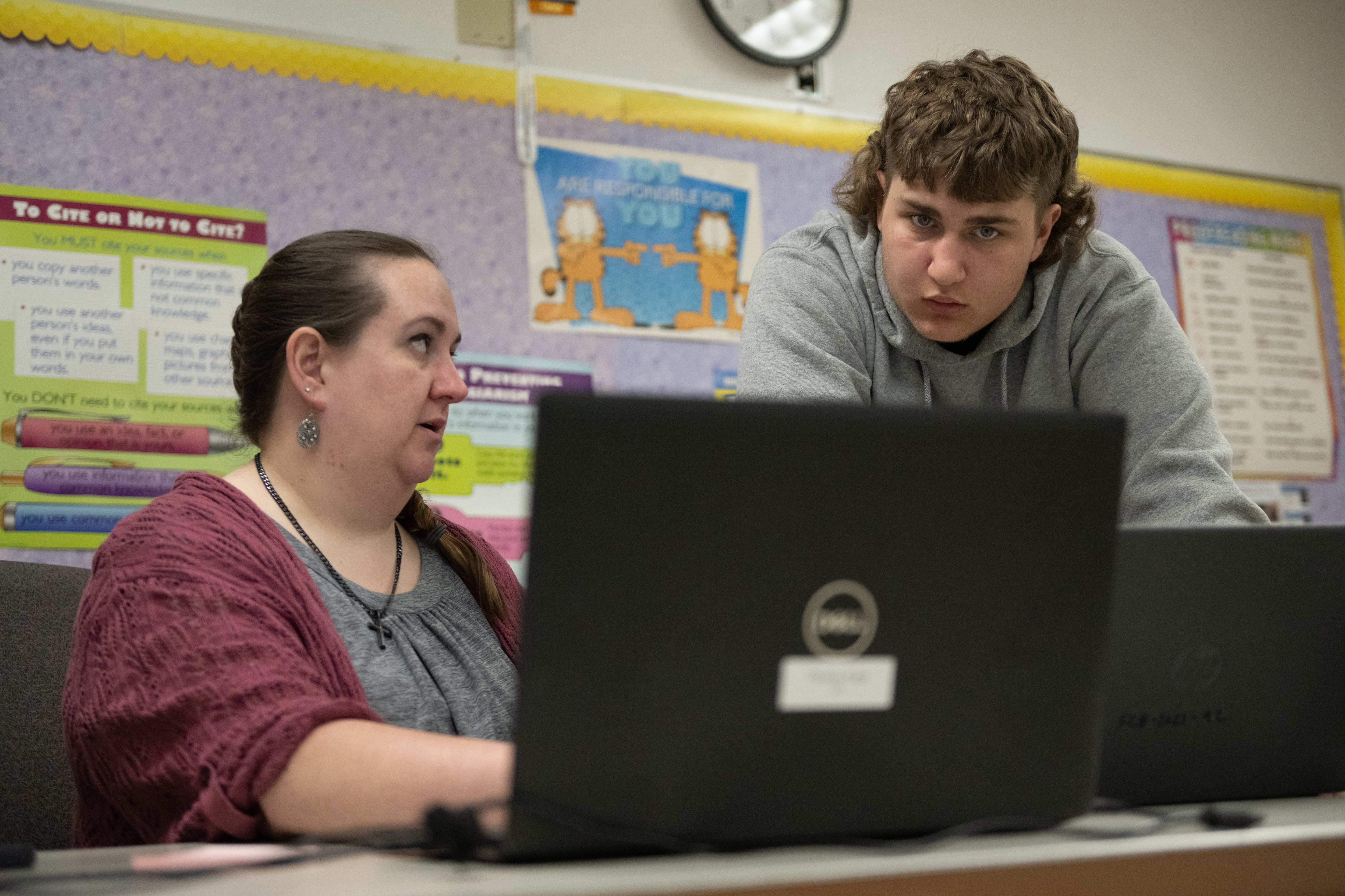 A woman in a cardigan sits at her desk in front of an open laptop. A young man or older teenager with floppy brown hair and a hoodie leans over the desk and looks at the screen. She appears to be explaining something to him.