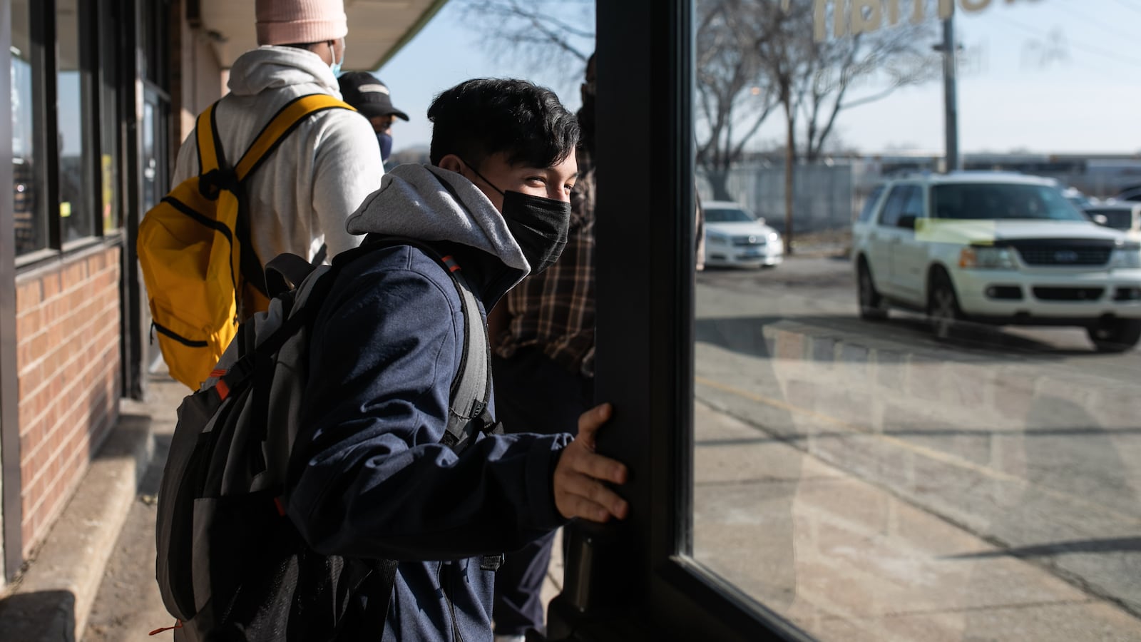 A student, wearing a black mask, blue jacket, and grey hoodie, holds a door open as he exits school. Other students are on the sidewalk directly behind him.