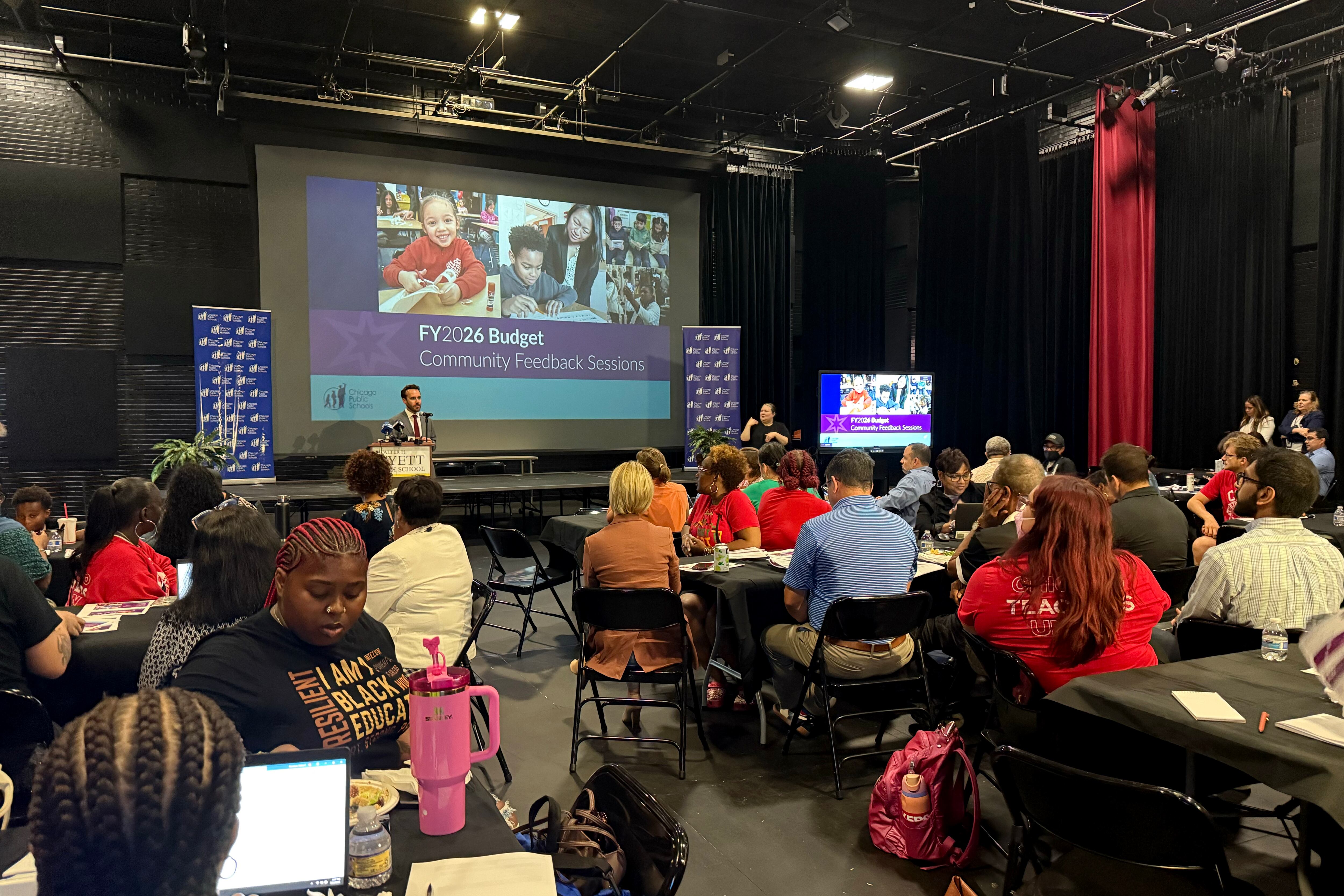 A room full of adults sitting in an auditorium facing a stage while one person stands at the front next to a large projector screen.