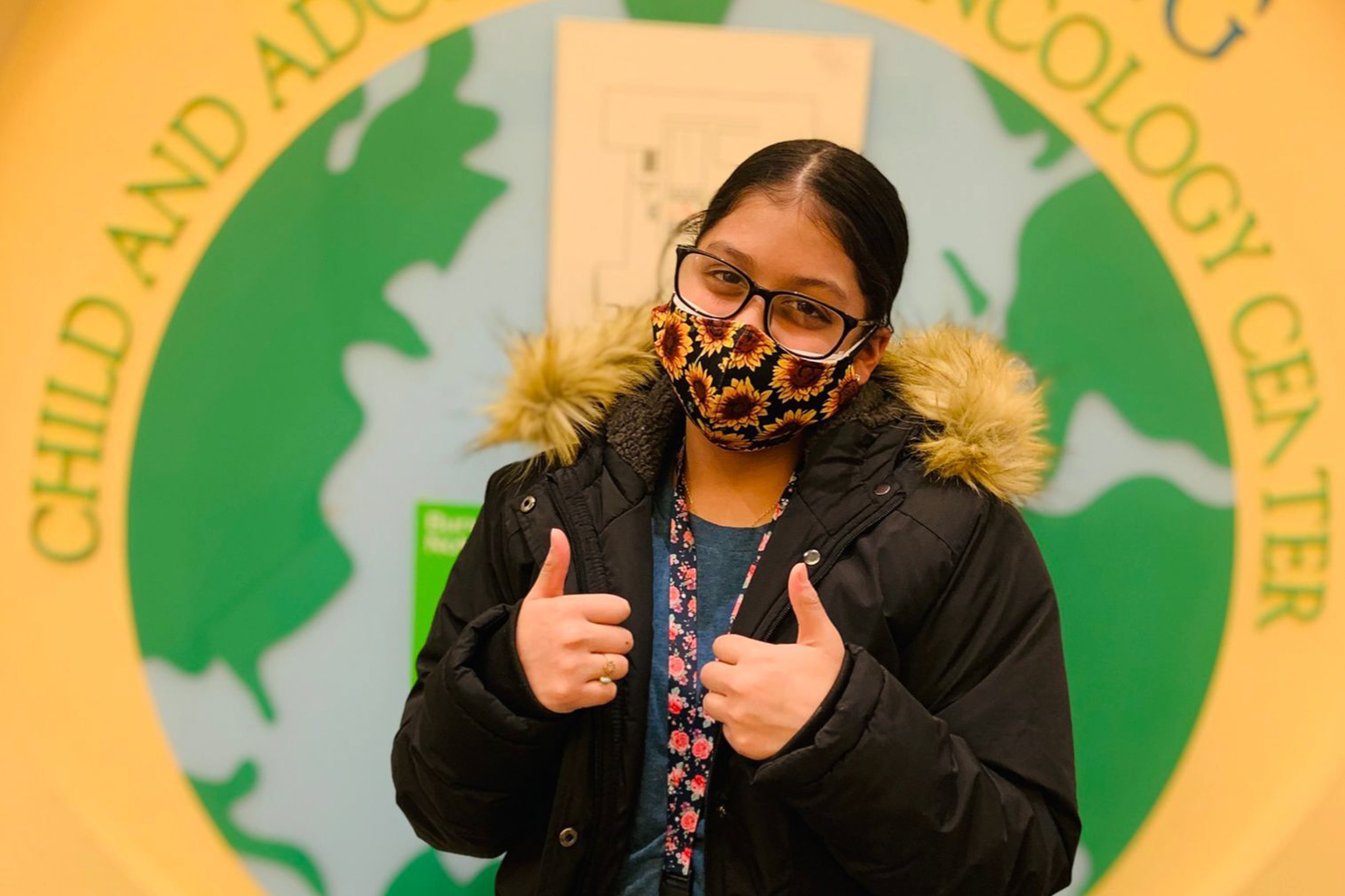 A young girl wearing a mask has two thumbs up in front of a sign.