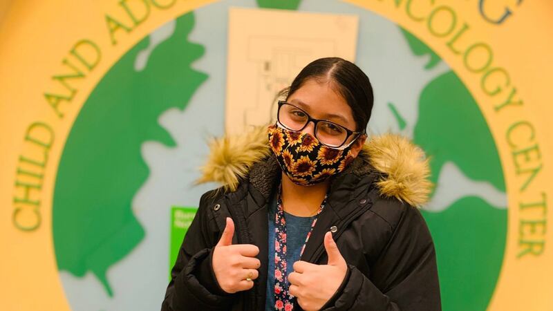 A young girl wearing a mask has two thumbs up in front of a sign.