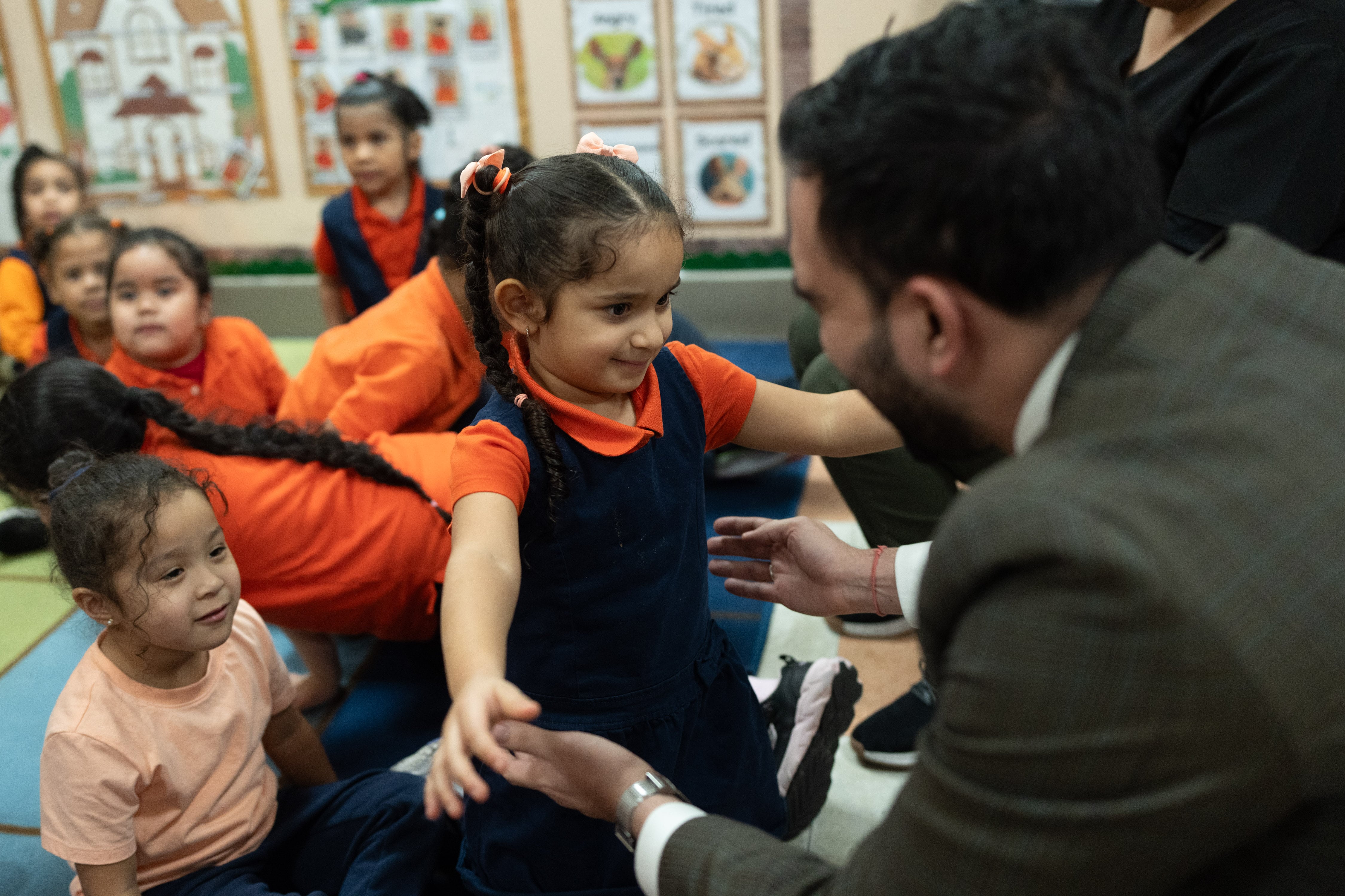 A man in a suit jacket holds his hands out to a little girl in a blue jumper and orange-red shirt.