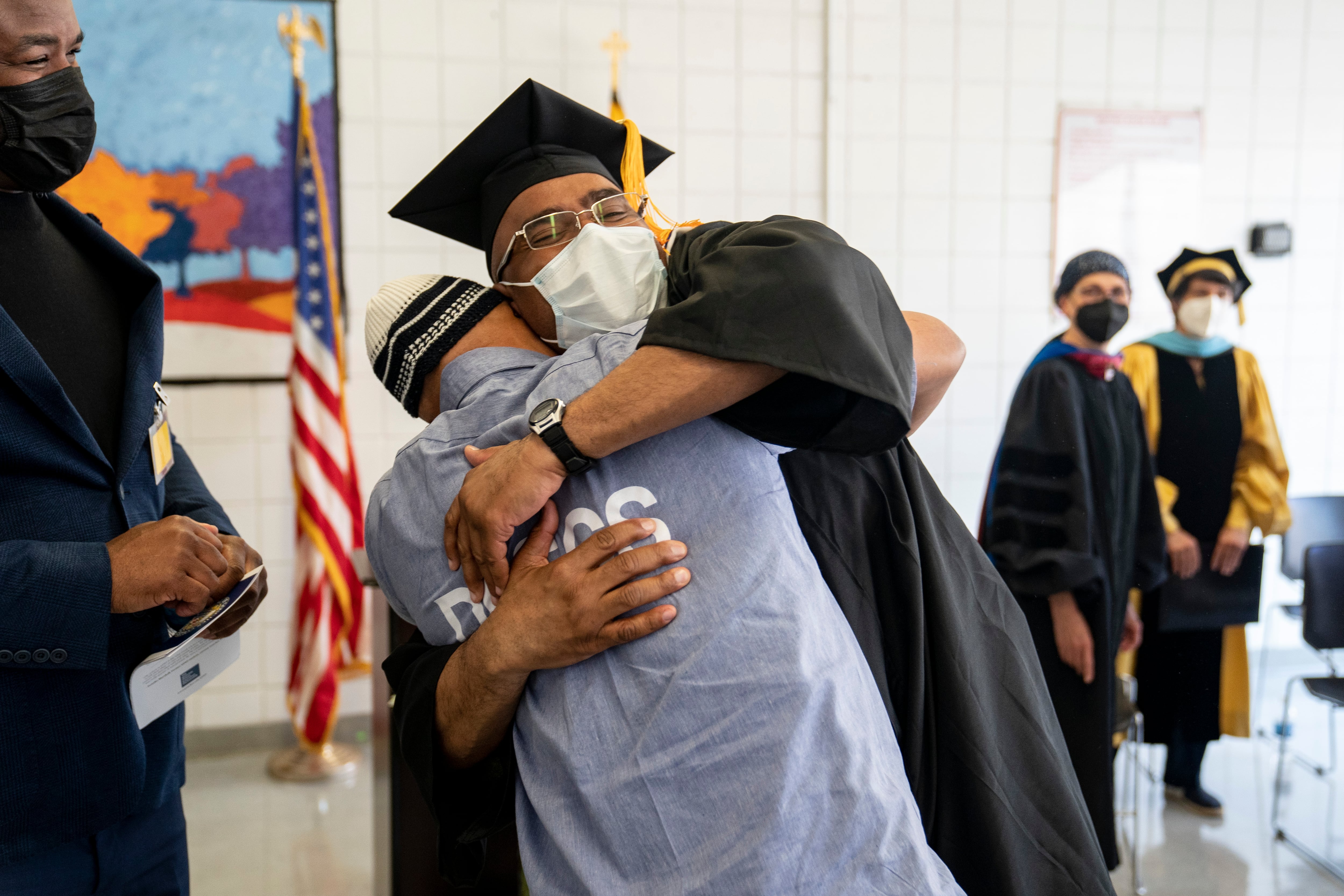 A man in a cap and gown hugs another man in a gray T-shirt and beanie in a room while three people look on. Everyone is wearing masks.