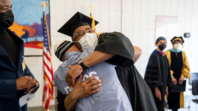 A man in a cap and gown hugs another man in a gray T-shirt and beanie in a room while three people look on. Everyone is wearing masks.