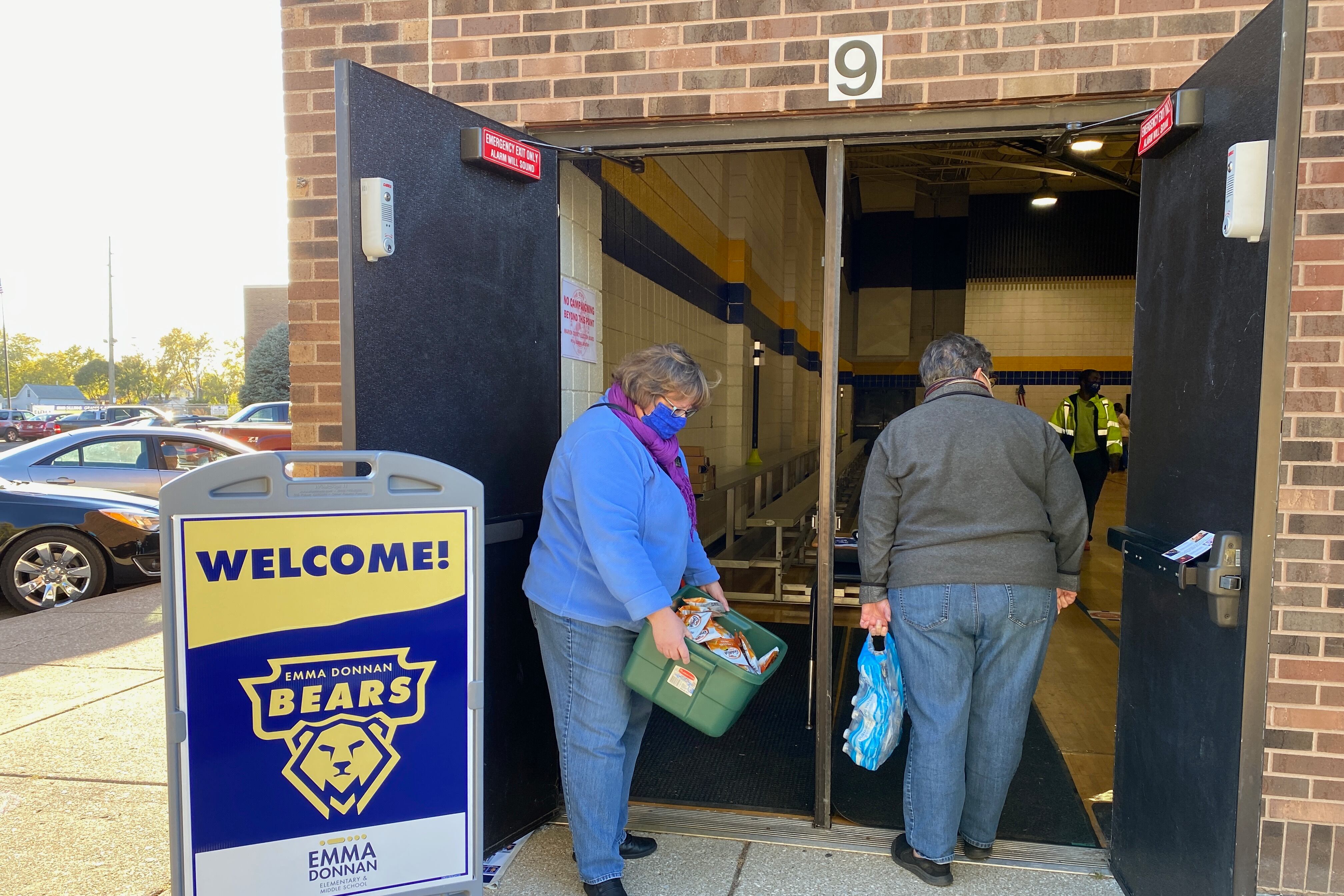 Two woman stand in the doorway of a polling place. One has a bin of snack and the other has little water bottles.