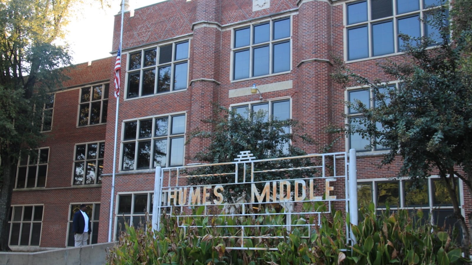 A metal sign that reads "Humes Middle" sits outside of a tall, red brick building with greenery and trees in the foreground and on the sides.