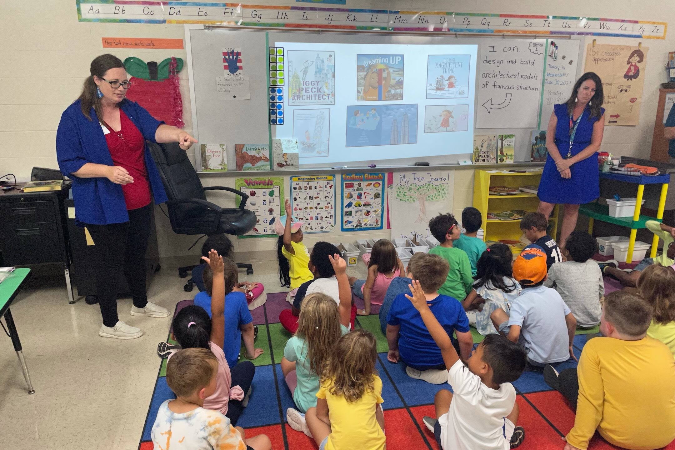 A dozen children sitting cross-legged on a multi-colored floor mat raise their hands to answer a question from one of two teachers standing before them in a classroom.