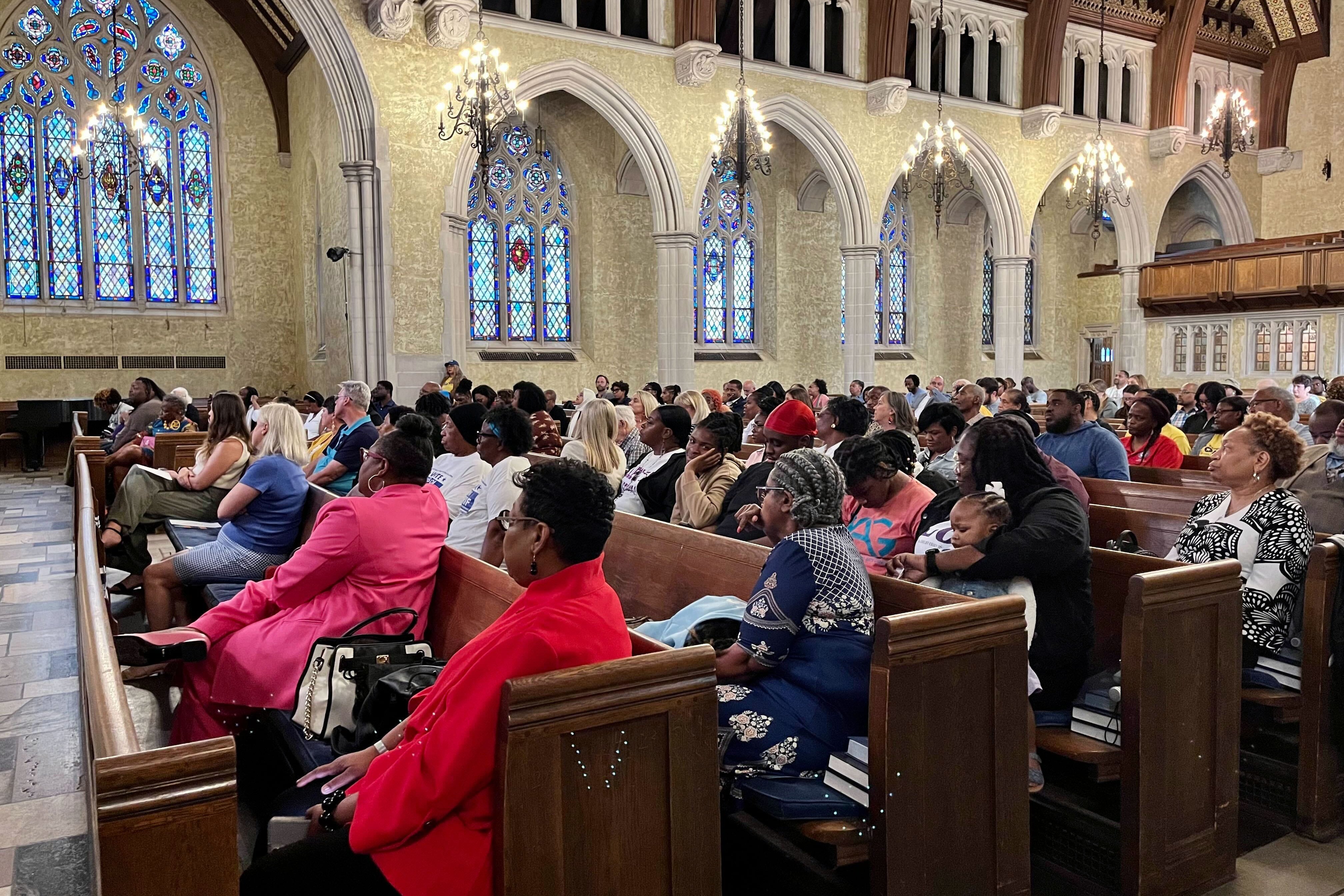 A large group of people sit in church pews with large arches and stained glass in the background.