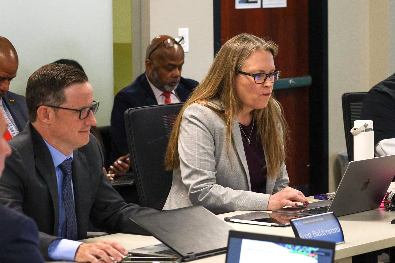 Two people sit at a table with laptops in front of them. There are two people in the background.