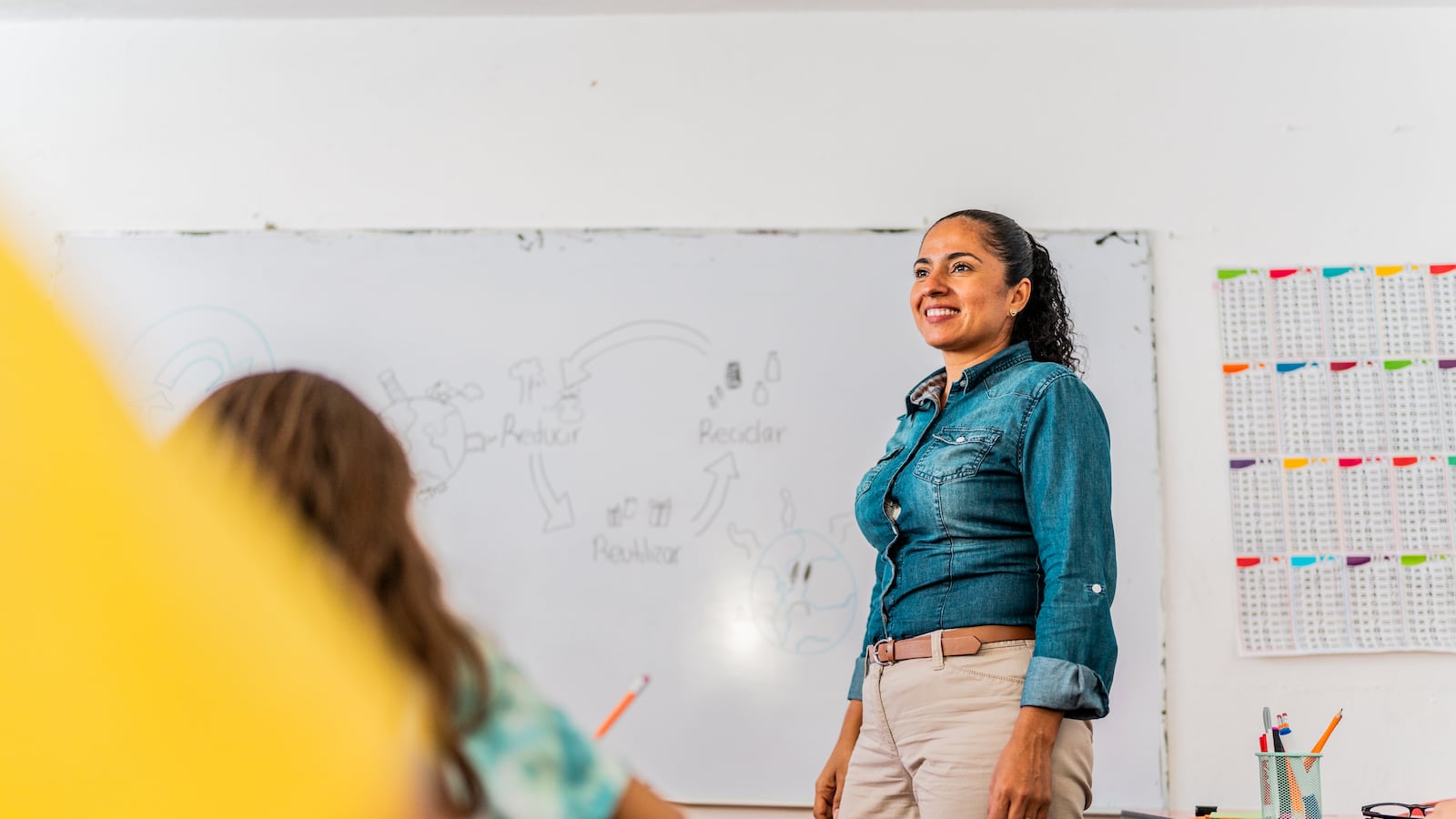 A woman in a blue denim shirt and khaki pants stands in front of a classroom.