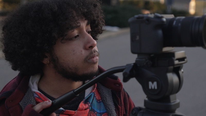 A teenage boy with dark hair stands behind a camera during a film shoot.