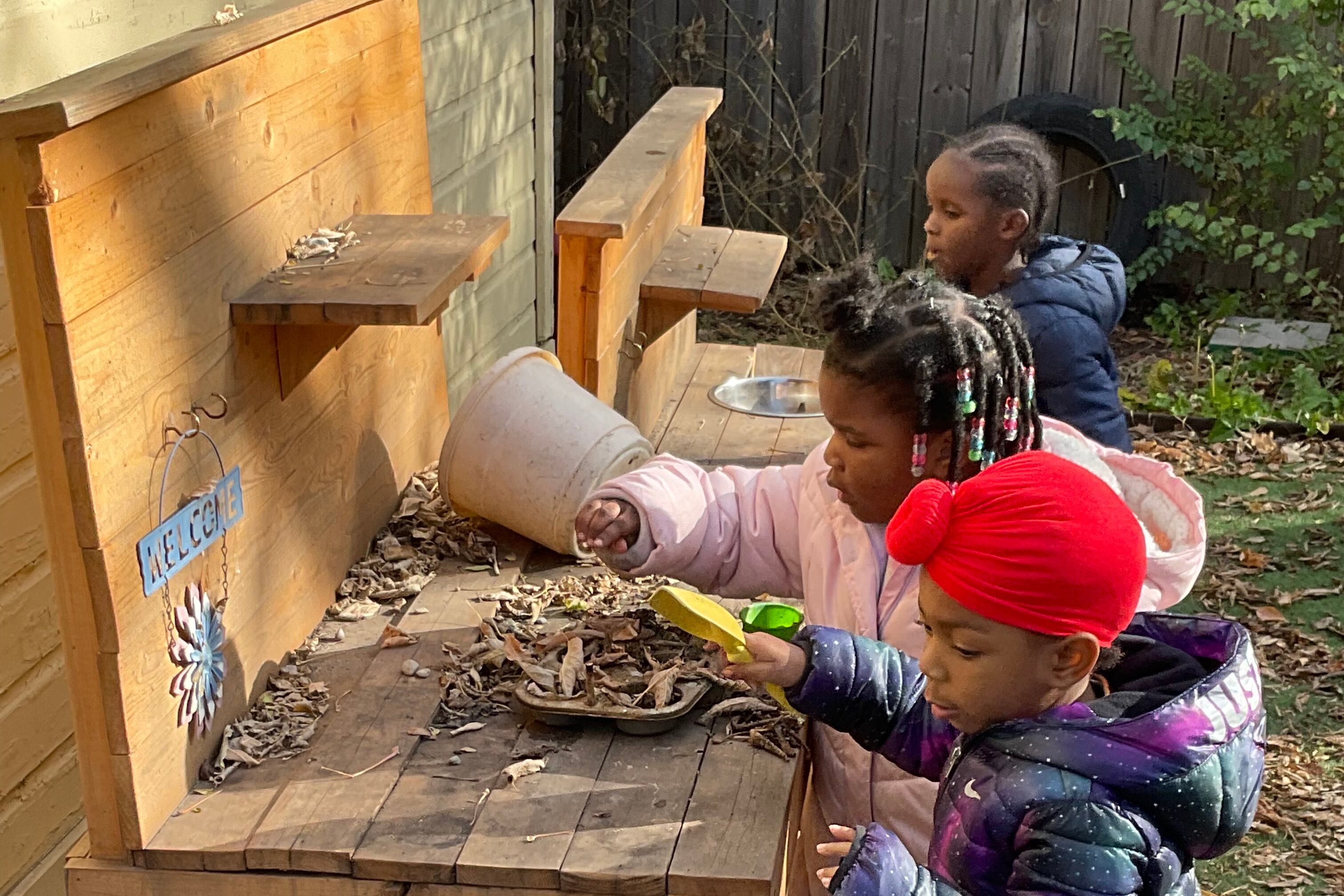 Children in coats pretend to cook at an outdoor play stove made out of wood using leaves as pretend food