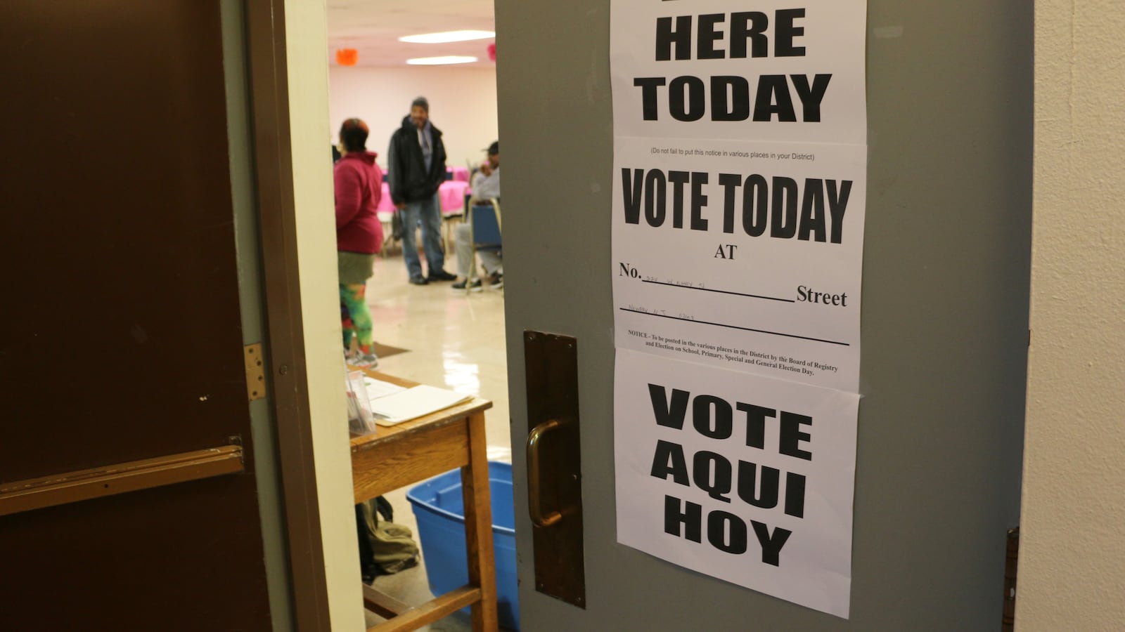 A door with white signs and black words that says "Vote here today. Vote today. and Vote aqui hoy." There are people in the background inside the room.