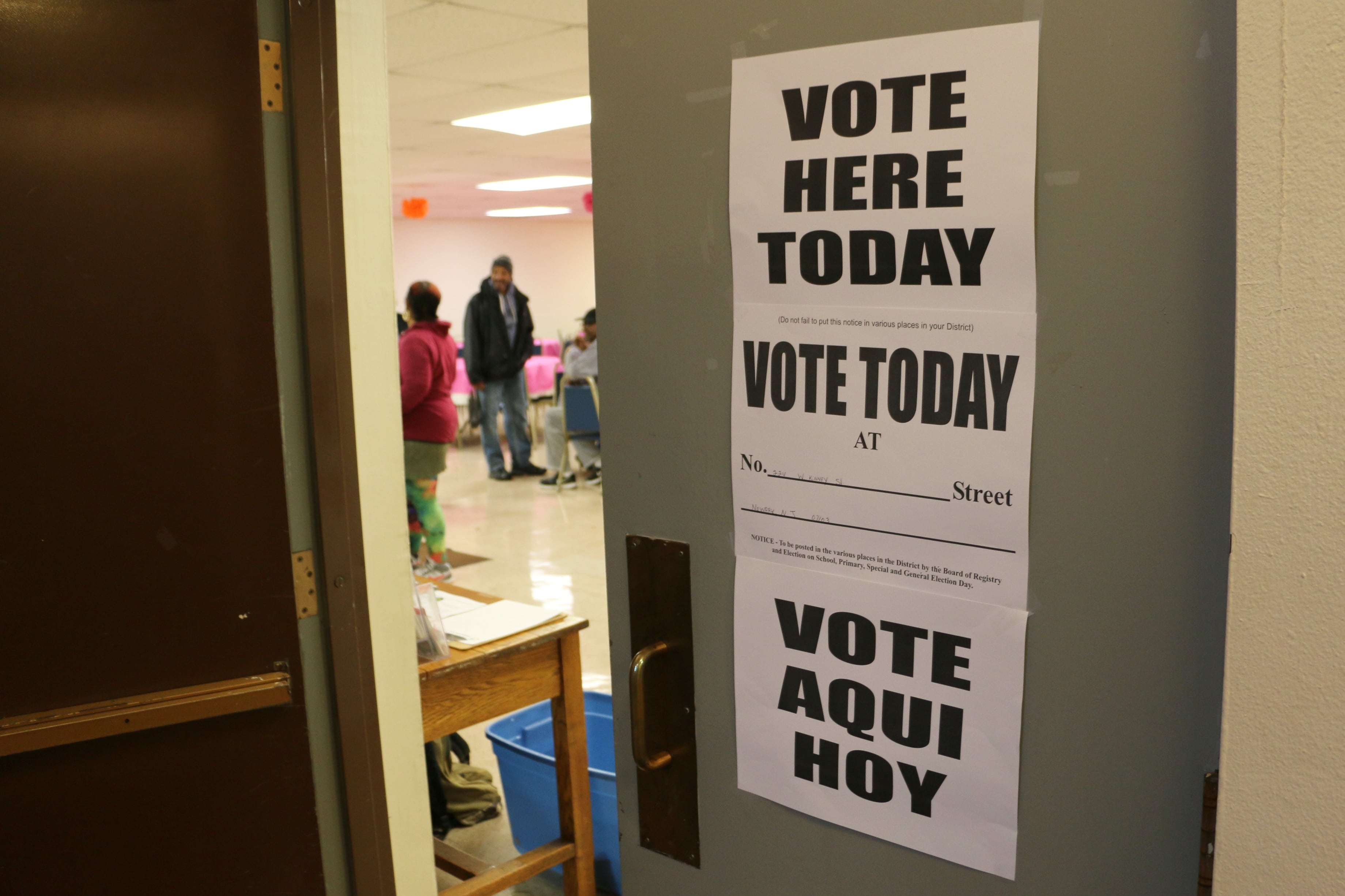 A door with white signs and black words that says "Vote here today. Vote today. and Vote aqui hoy." There are people in the background inside the room.