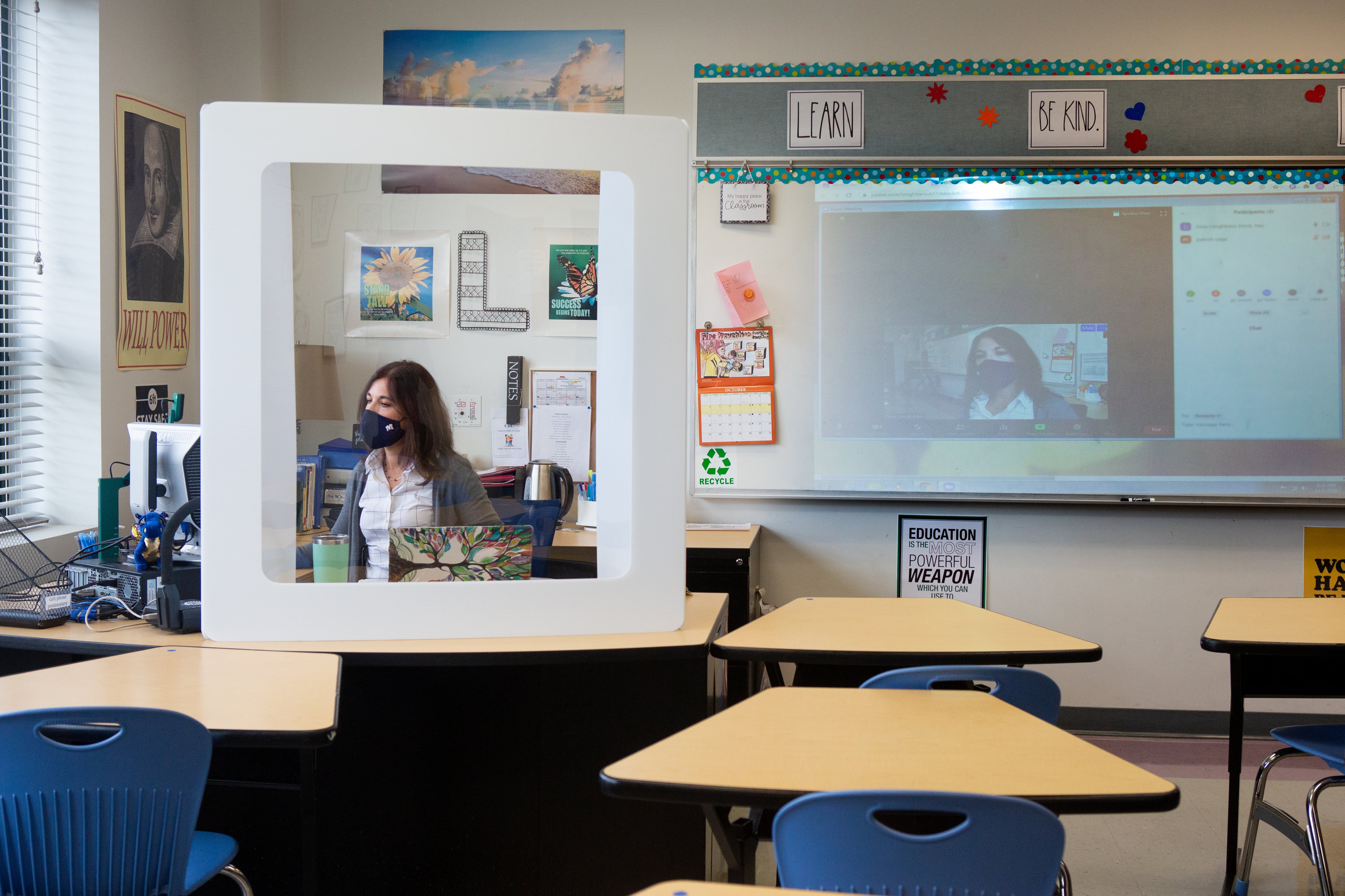 Teacher wearing a face mask seated behind a desk shield.