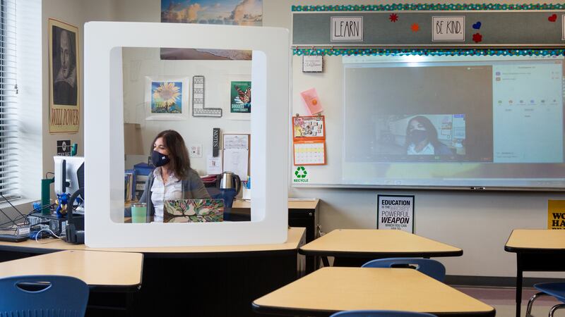 Teacher wearing a face mask seated behind a desk shield.