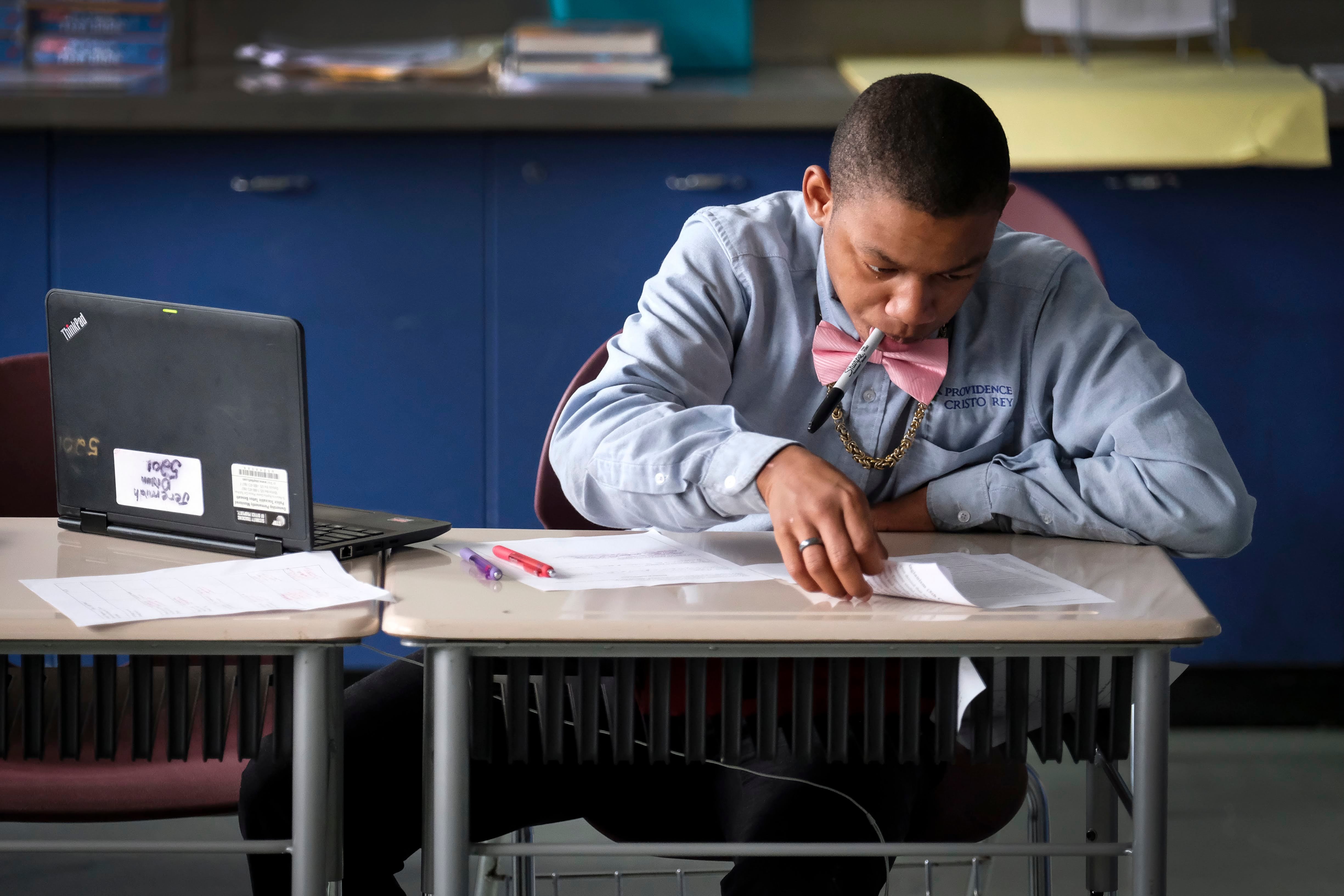 A photograph of a Black high school boy sits at his desk holding a black sharpie in his mouth while working on classwork.