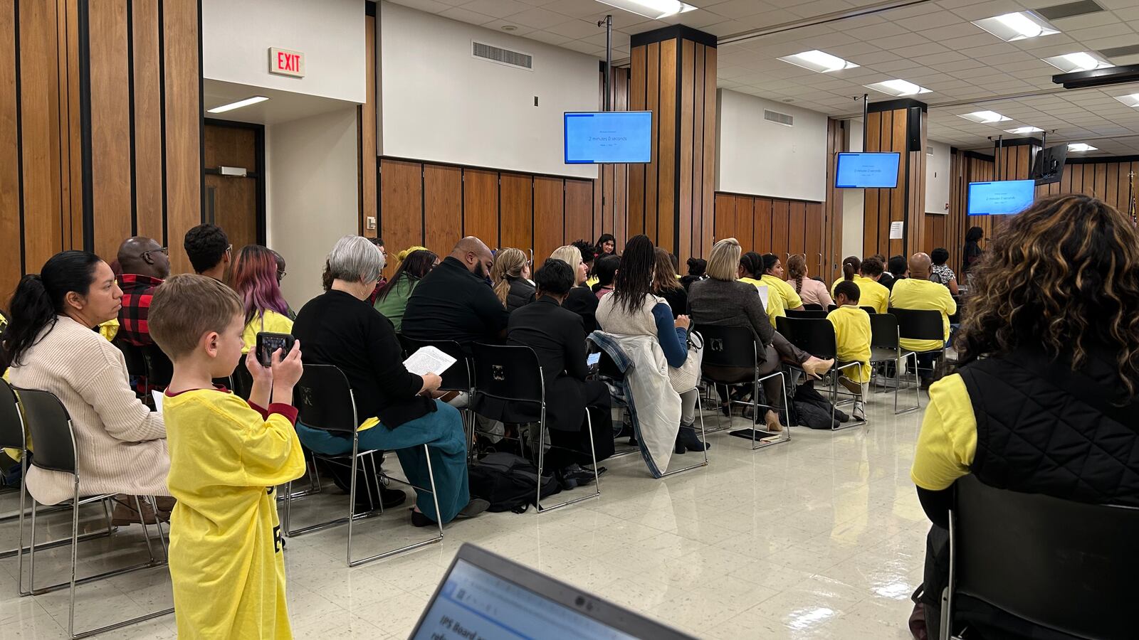 A child in a yellow shirt stands in an aisle during a public meeting. He holds a phone to take a picture or record or a video.