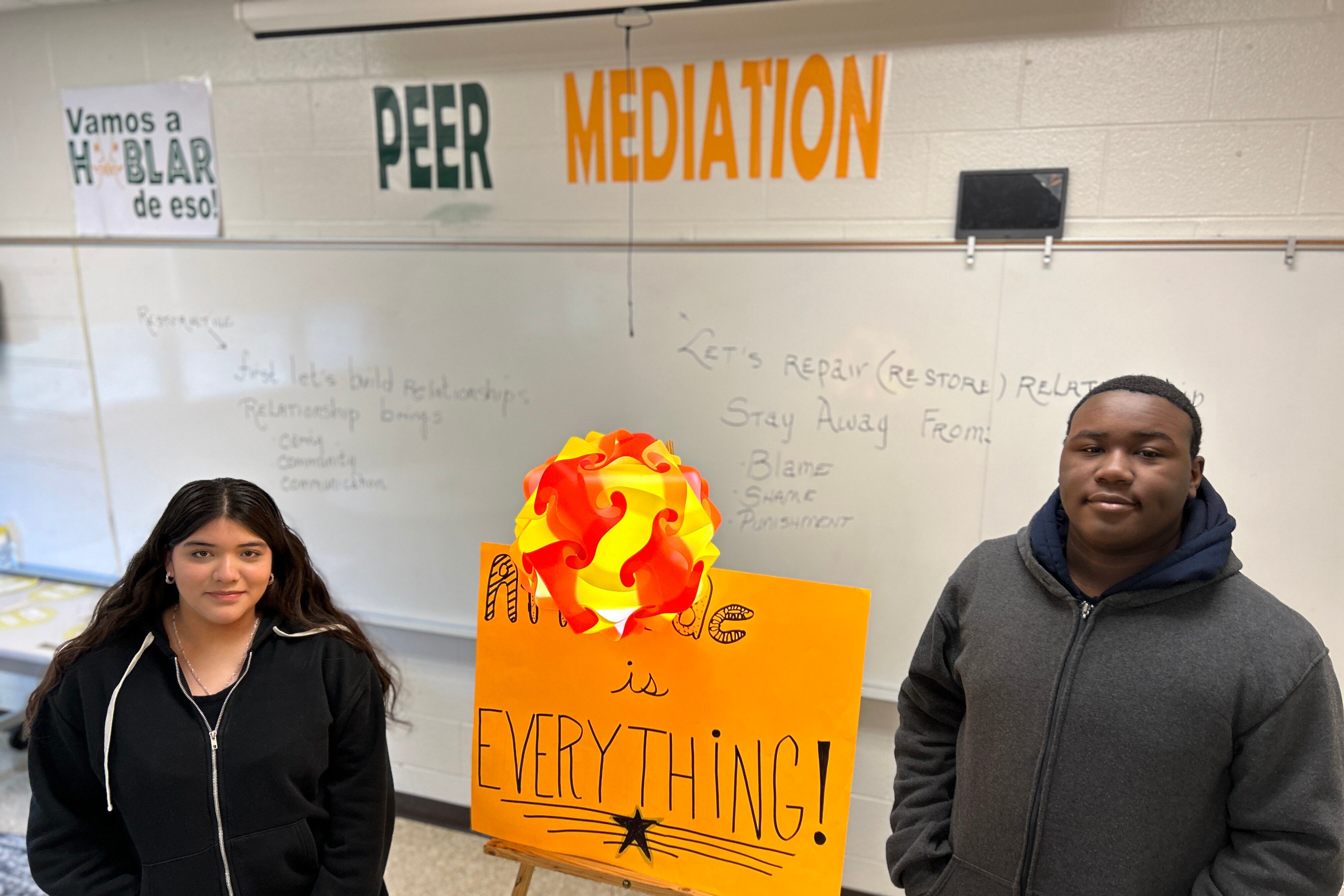 Two students both wearing zip up sweaters stand on each side of an orange poster board that is standing up in front of a dry erase board in a classroom.