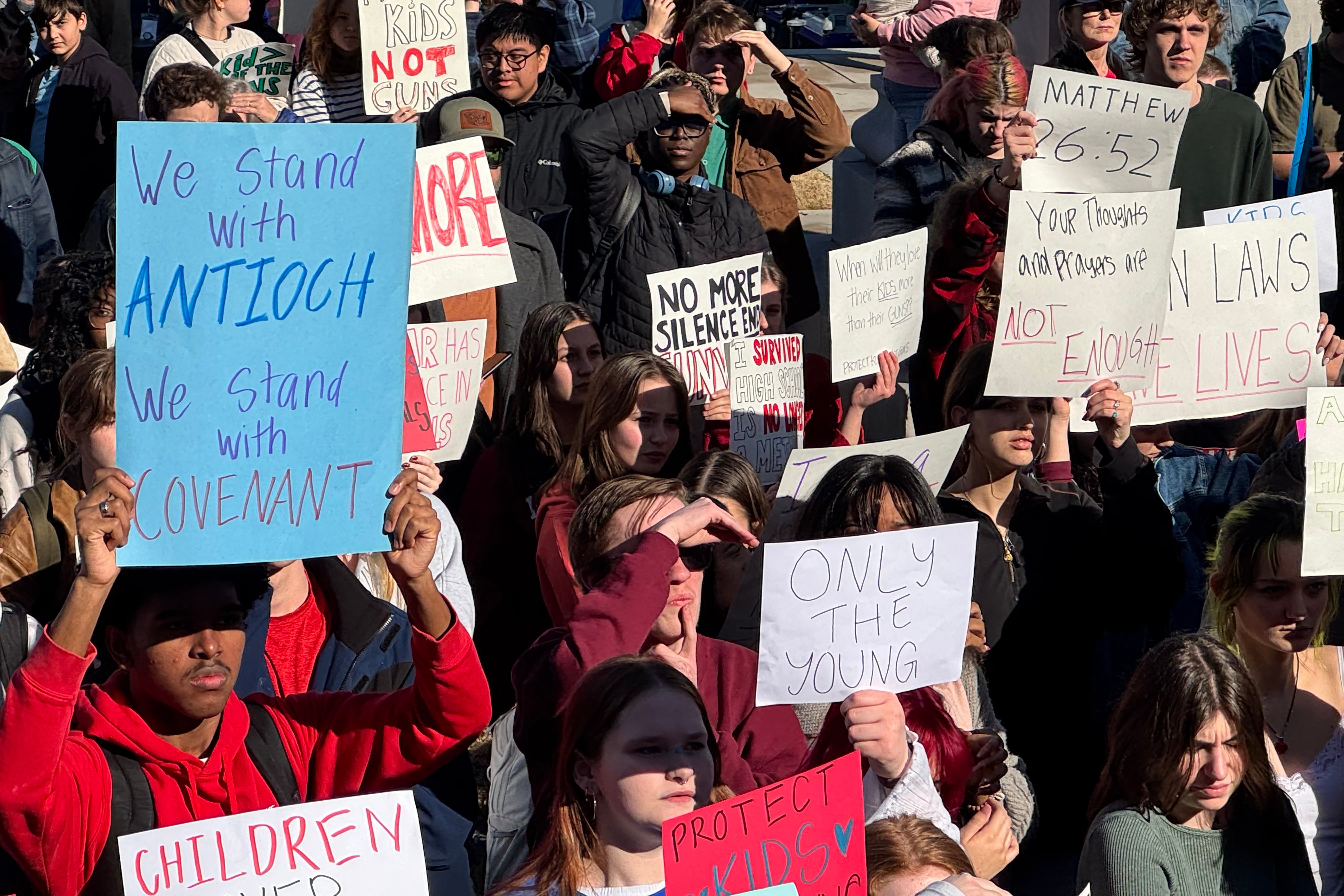 A large crowd of people, many are holding signs, protest outside on a sunny day.