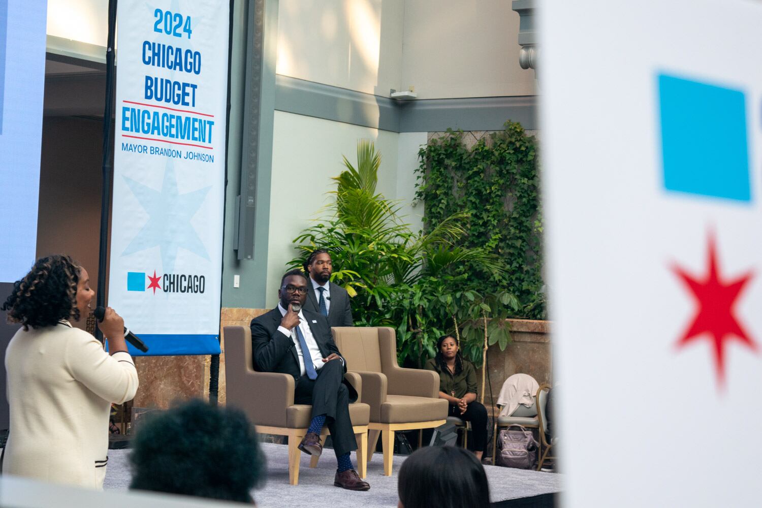Mayor Brandon Johnson sits in on a chair on a stage. He is wearing a suit and tie and has his finger touching his face as he looks toward Annette Guzman, who is off the stage and to his left. Guzman is the city’s budget director. She is speaking into a handheld microphone. A sign on the stage says “2024 Chicago Budget Engagement.” In the foreground is a blurred white sign with the Chicago red star and a blue square above it.