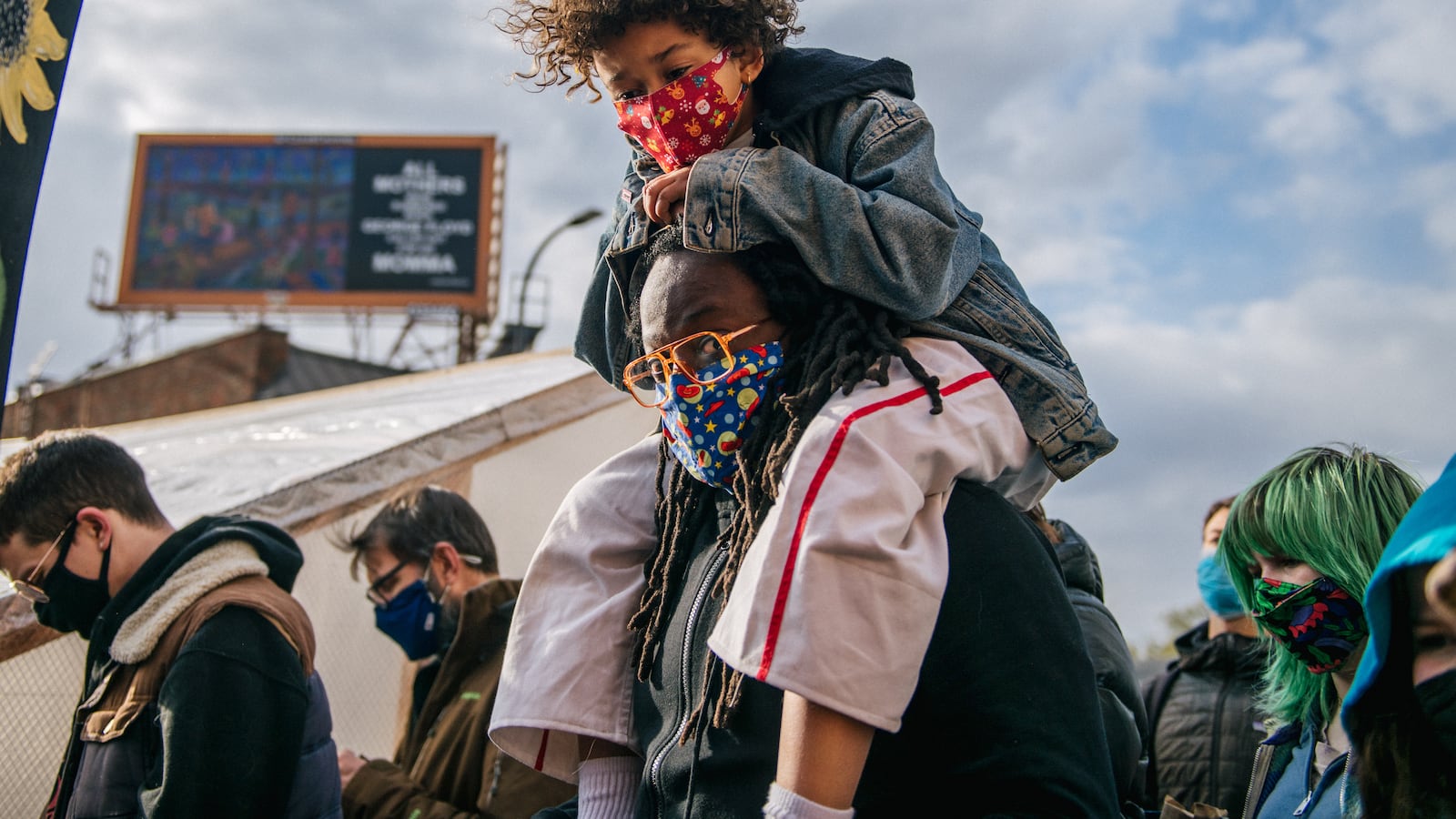 A child wearing a red mask, jean jacket and white pants with a red stripe is carried on their father’s shoulders, who wears orange-rimmed glasses and a blue mask with designs. Many people around them are bowing their heads.