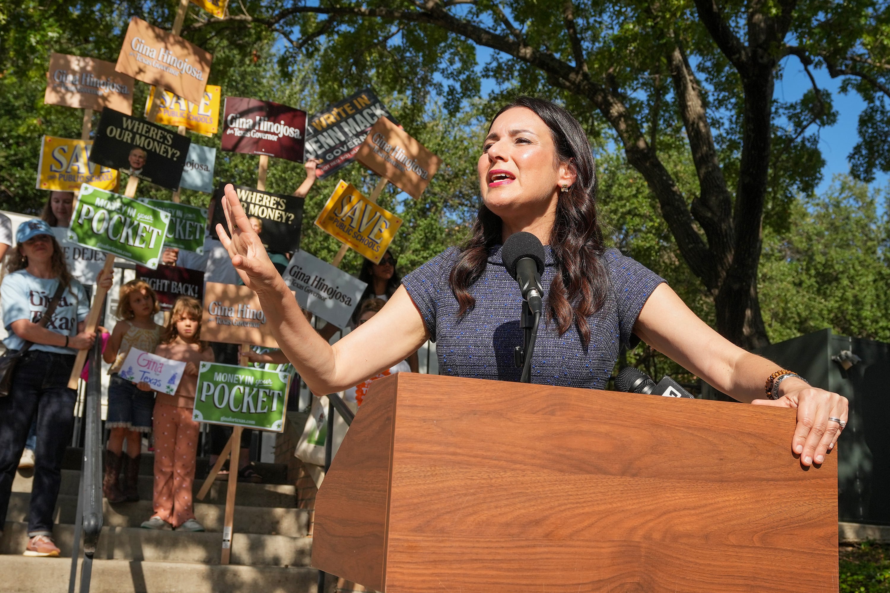 A woman speaks from behind a wooden podium with a group of people holding signs in the background on a sunny day.
