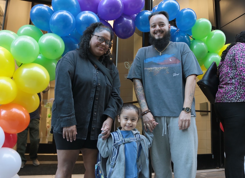 Two adults stand holding their young son's hands outside of a stone building and in front of an entrance with a colorful balloon arch.