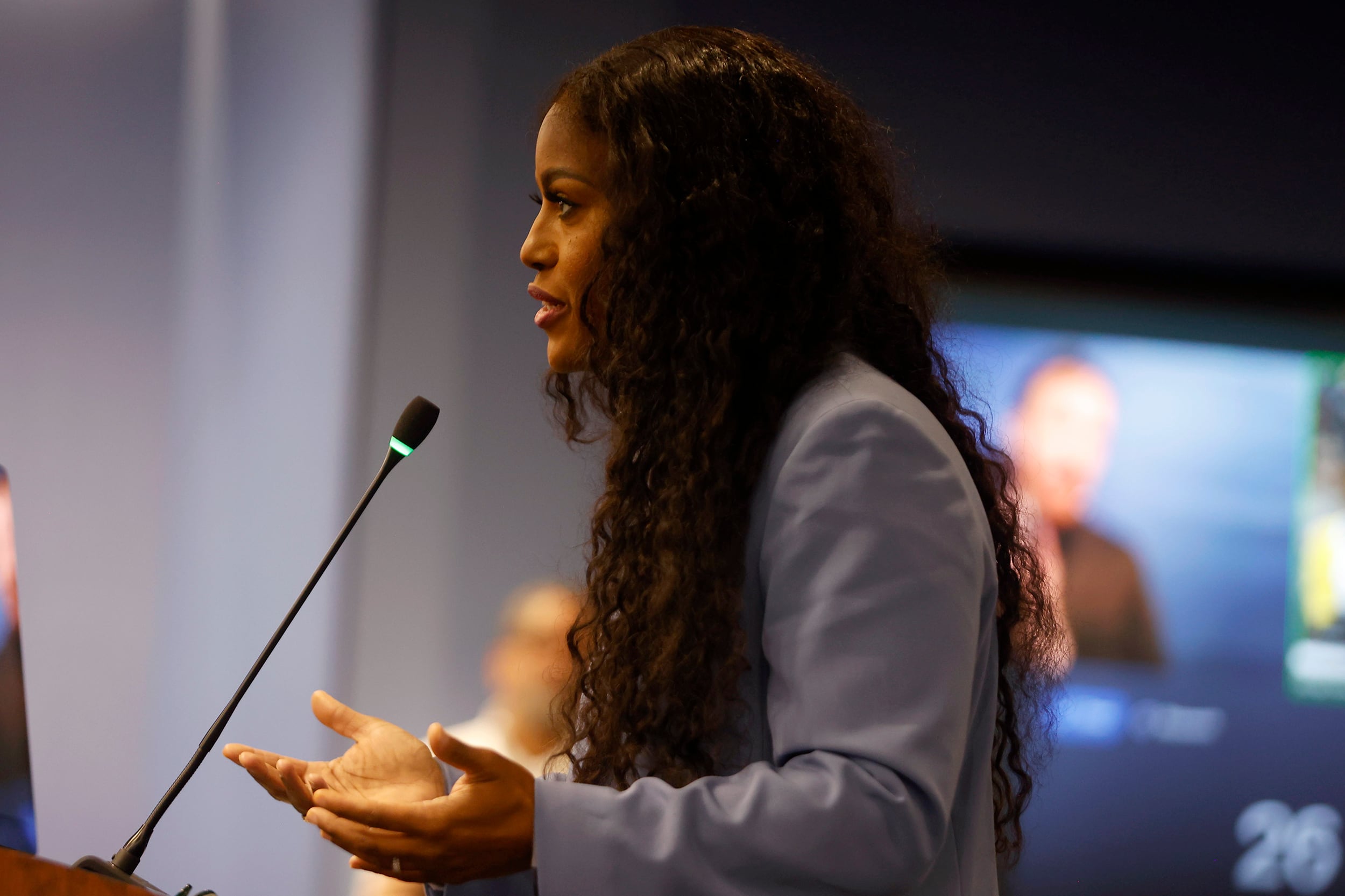 A Black woman with long, dark and curly hair and wearing a blue suit speaks from behind a podium and in front of a crowd.