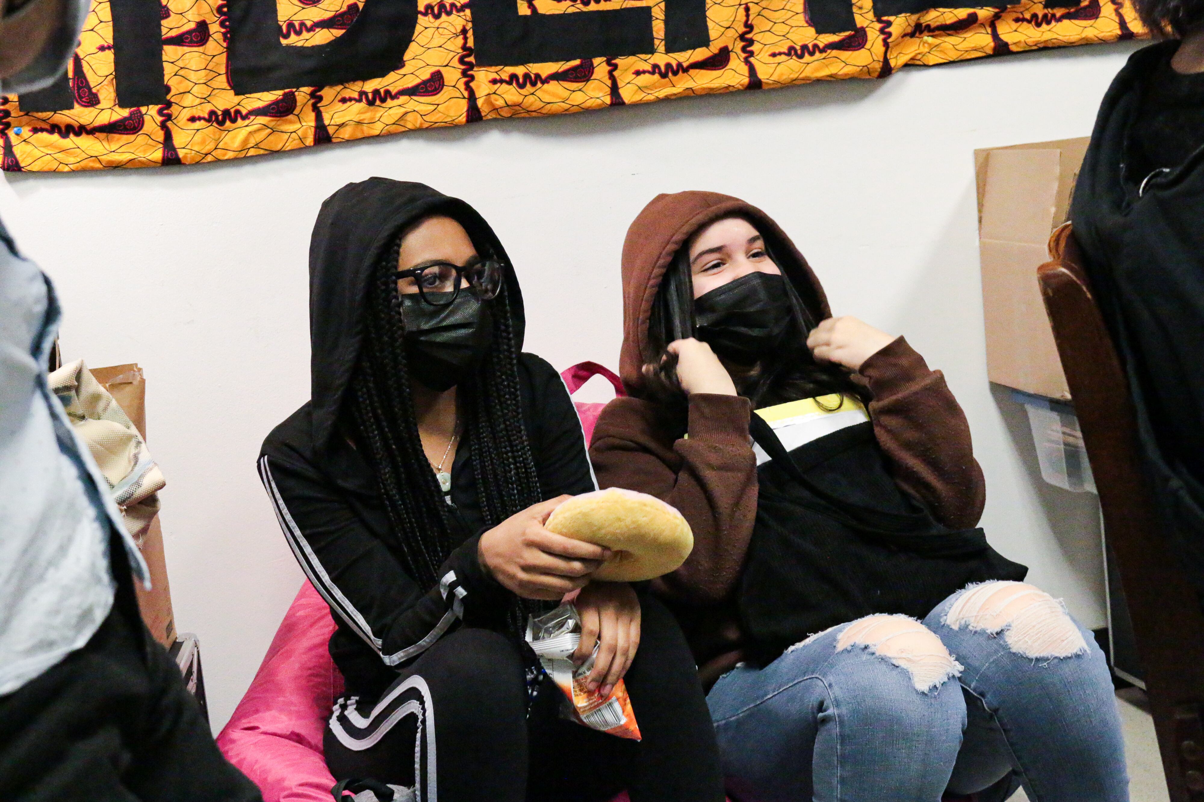 Two students in masks and hoodies lean against a wall under a banner.