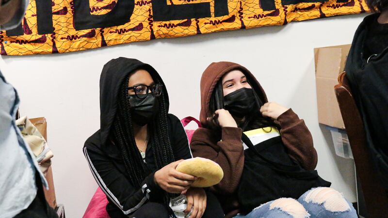 Two students in masks and hoodies lean against a wall under a banner.