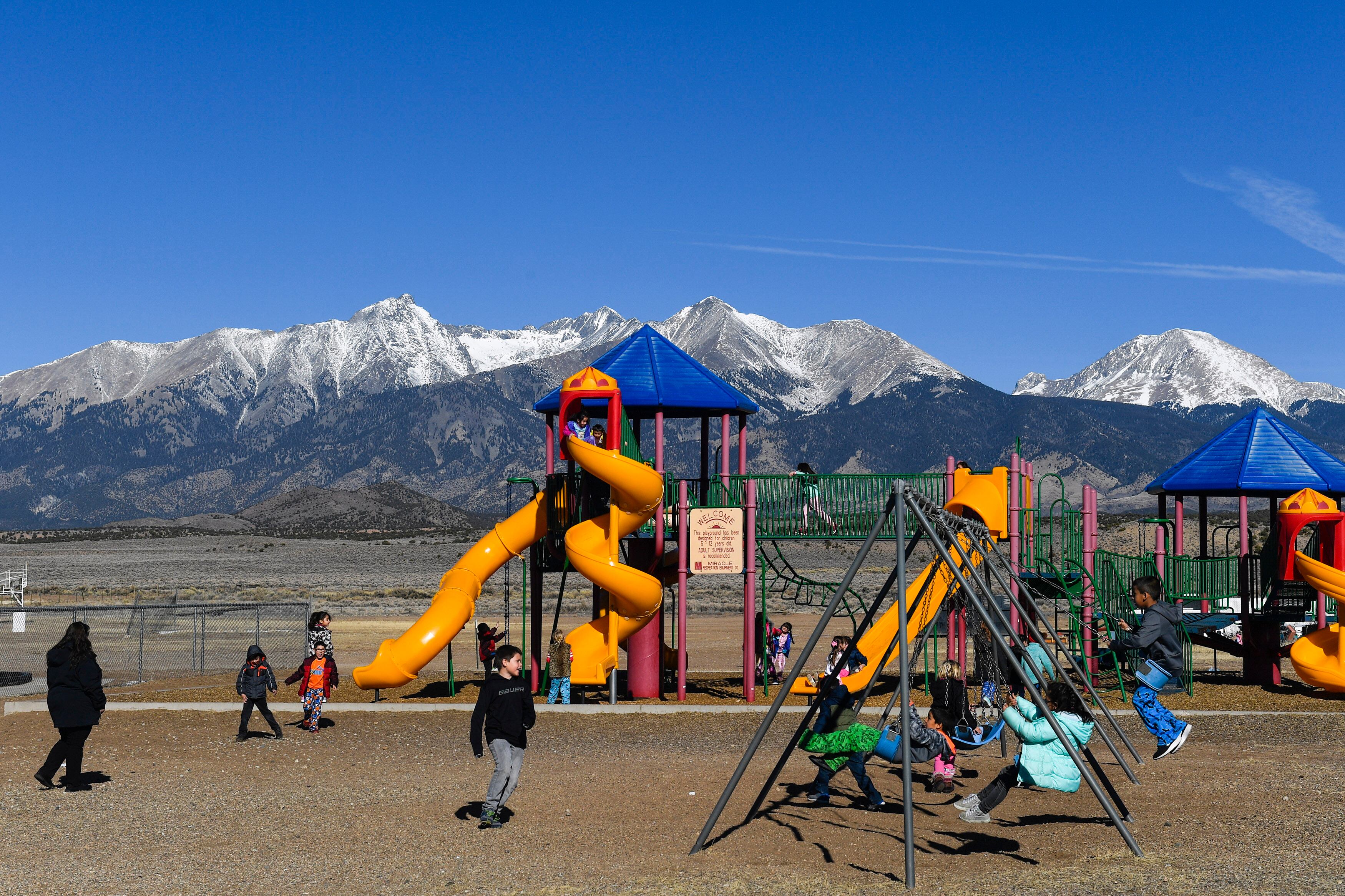 Young students play on a colorful playground with large mountains and a blue sky in the background.