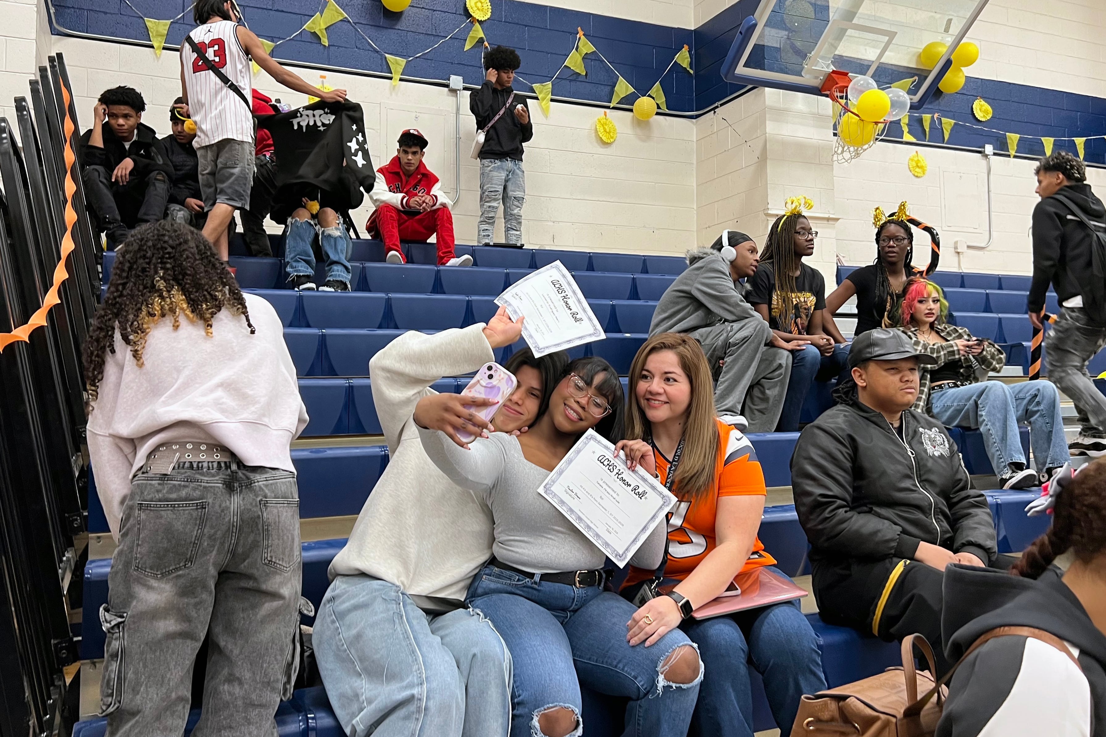 A photograph of a high school gymnasium full of high school students sitting in the bleachers.